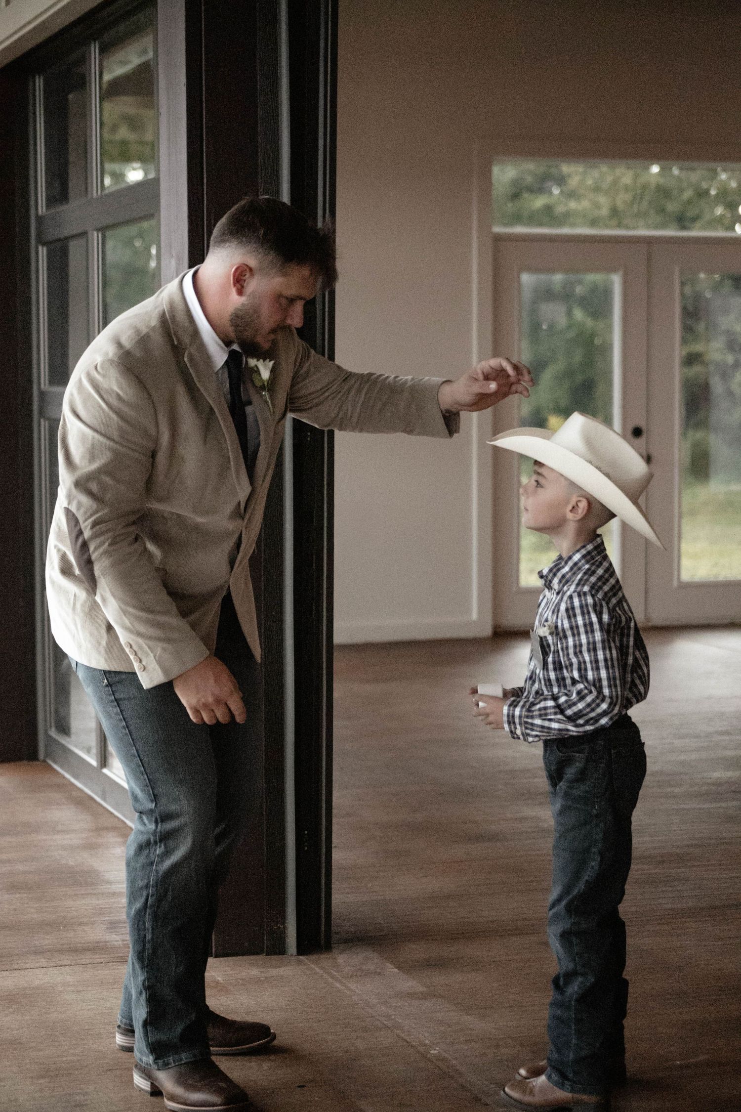 Man in tan blazer interacts with young boy wearing cowboy hat and plaid shirt indoors.