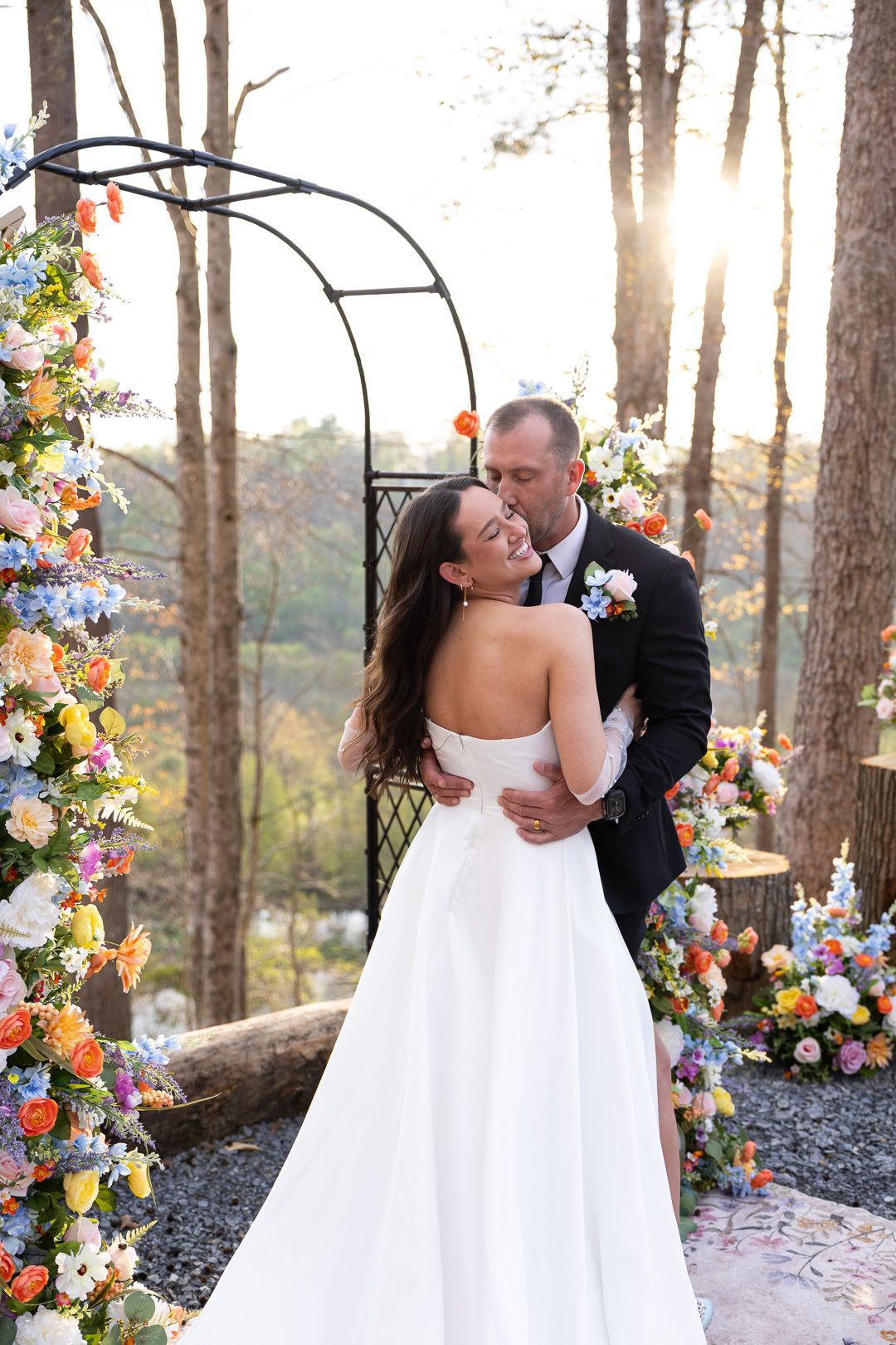 Newlyweds embrace under a sunlit floral arch with vibrant colorful flowers at outdoor wedding ceremony.