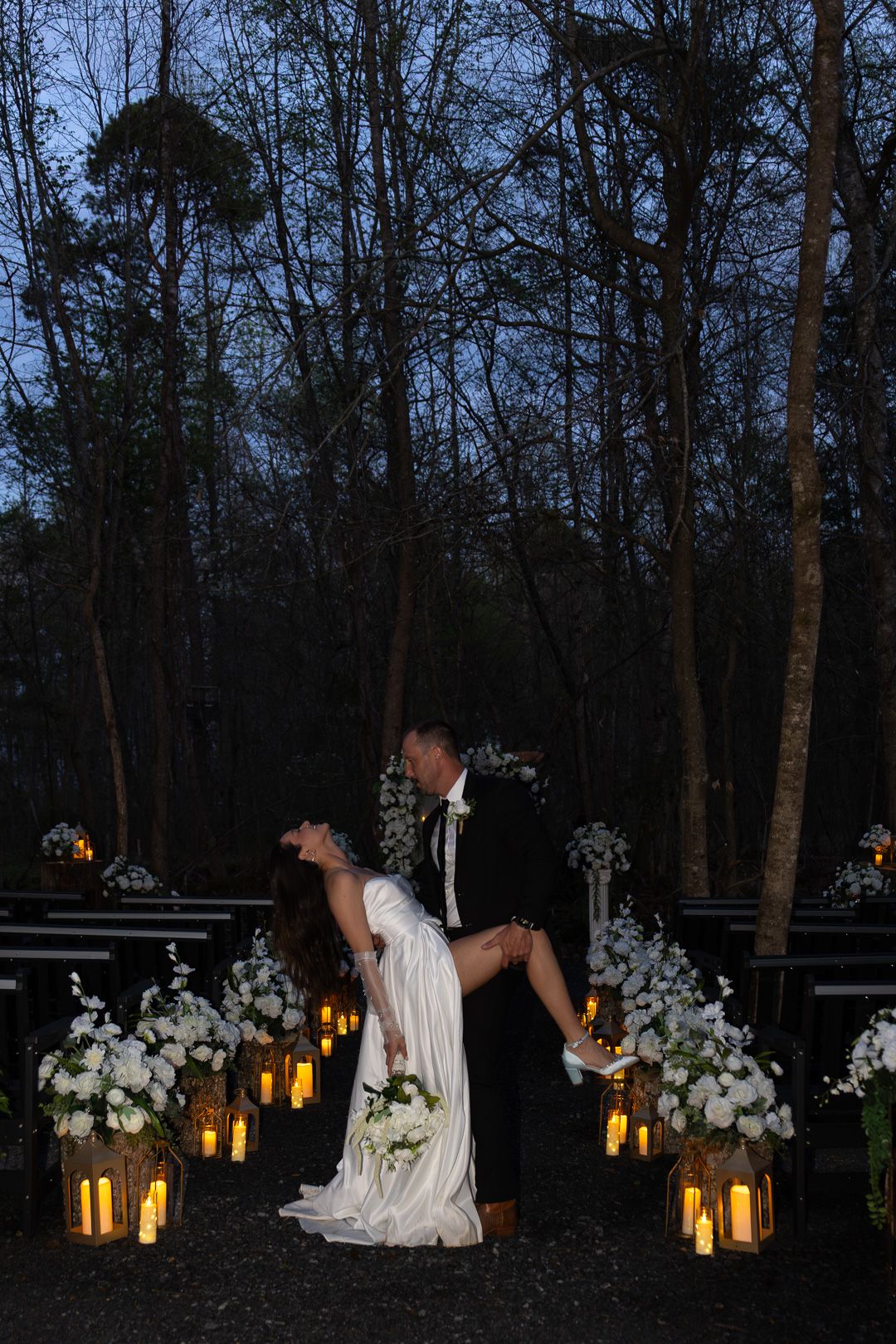 Groom lifts bride in a romantic dip along a candlelit forest wedding aisle at dusk.