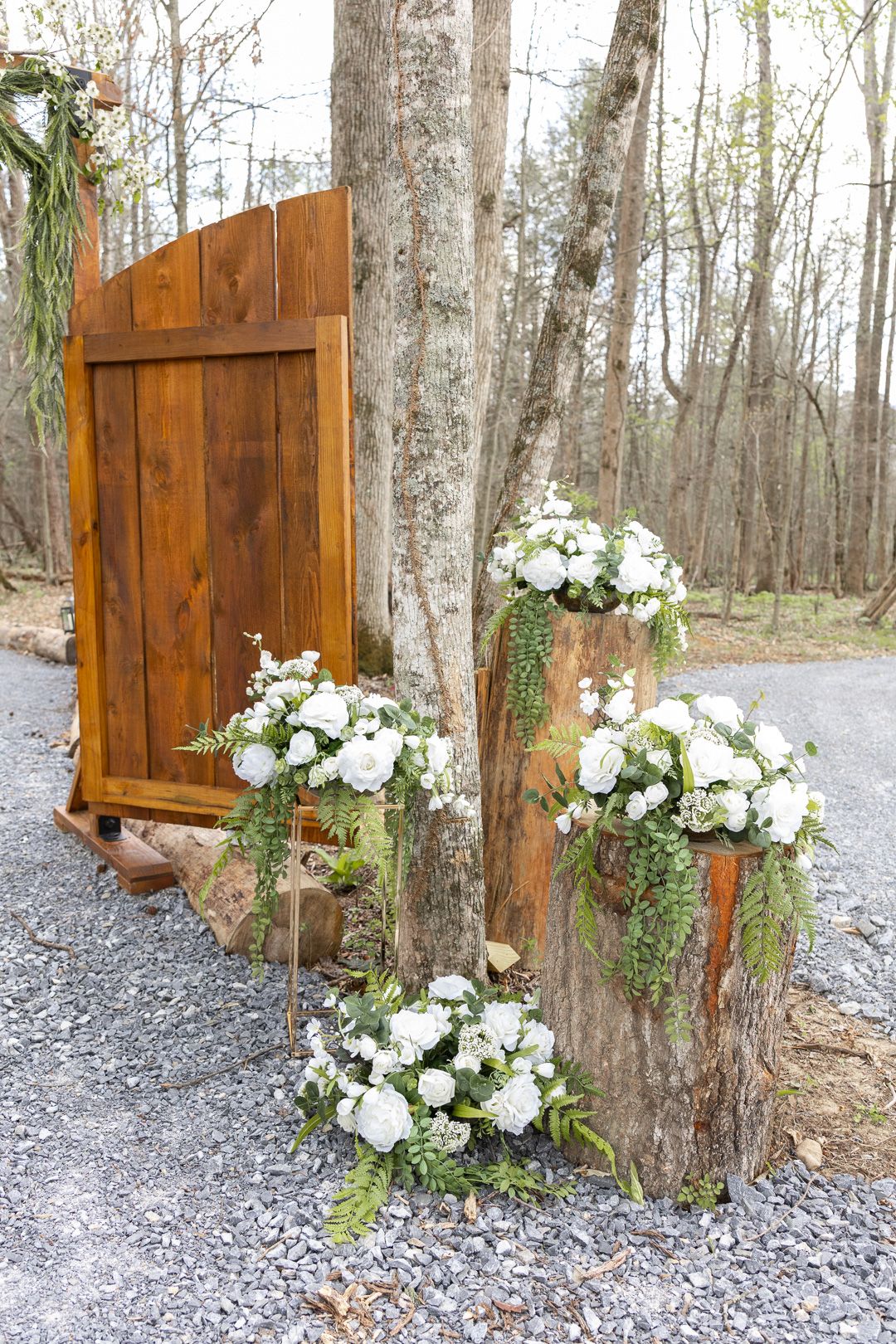 Rustic wooden door wedding entrance with white floral arrangements on tree stumps in forest setting.