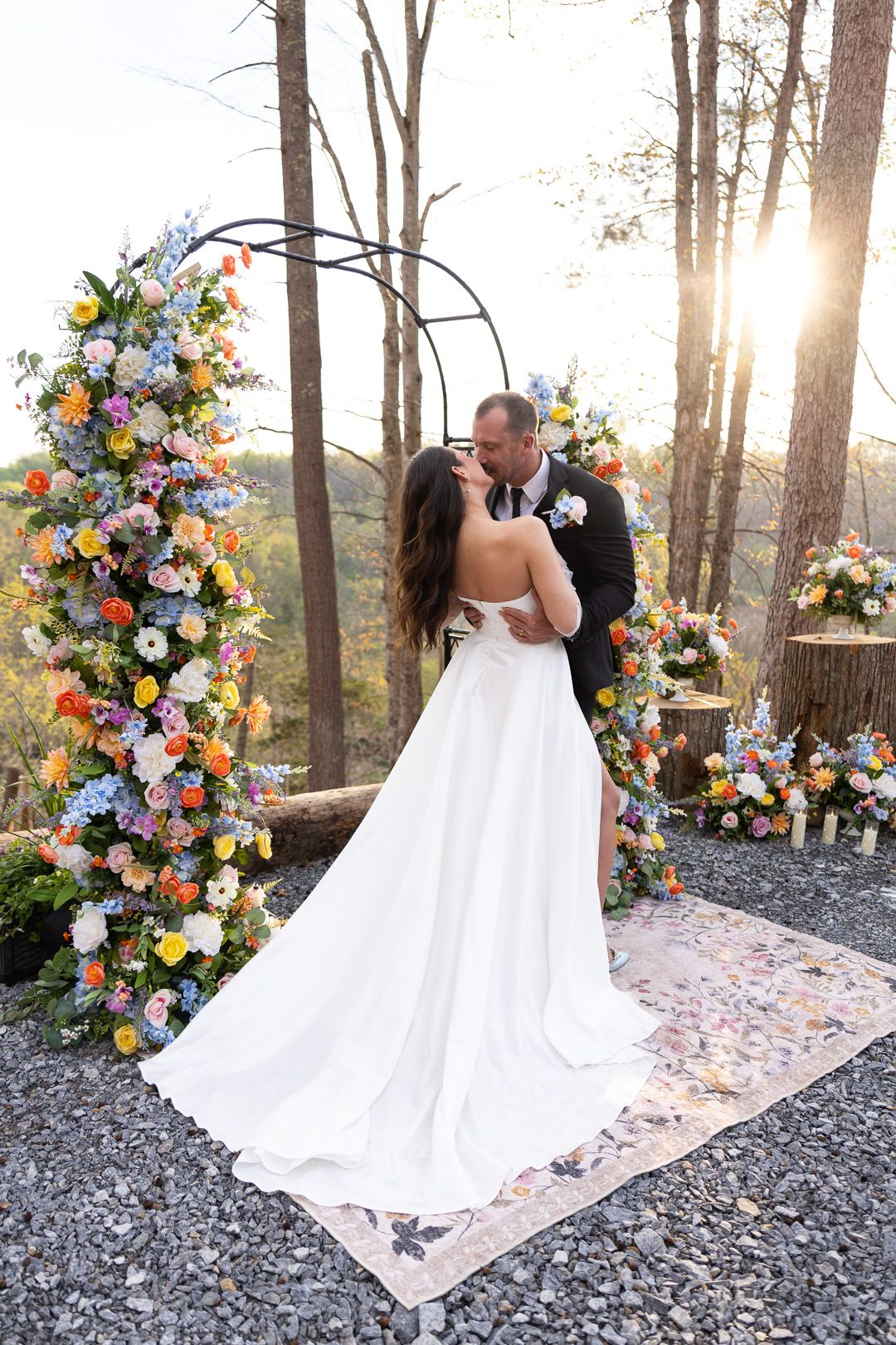 Bride and groom share first kiss under colorful floral arch at outdoor wedding ceremony in wooded setting at sunset.