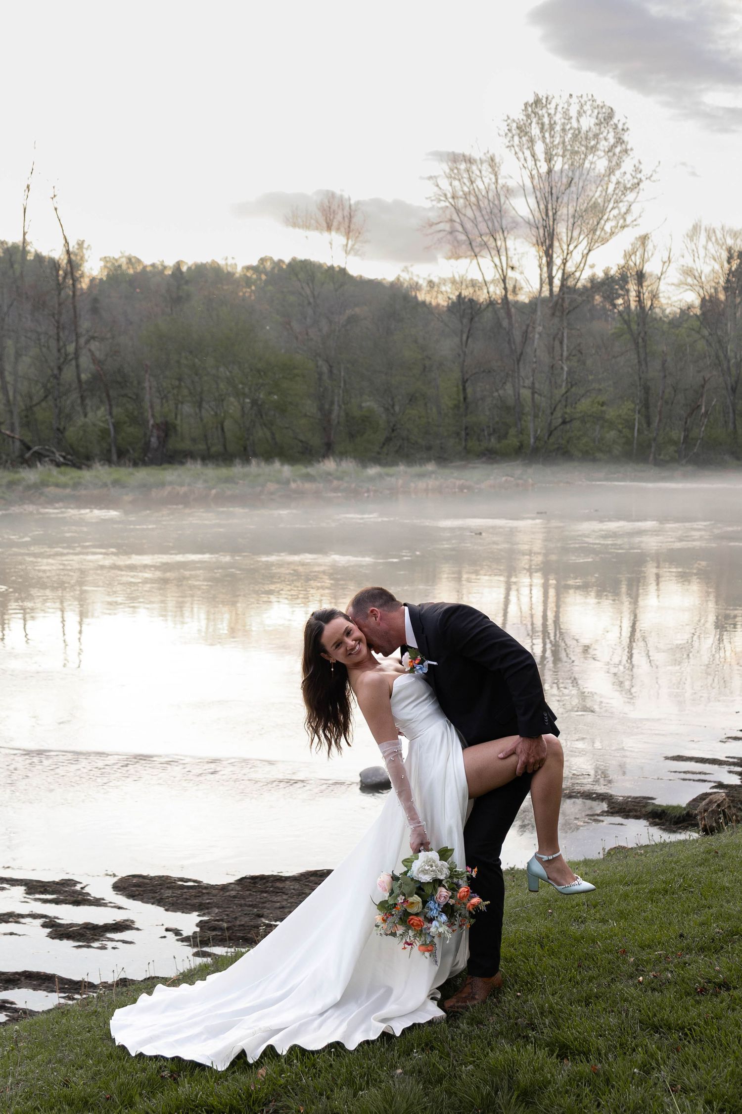 Bride and groom share a romantic dip kiss by a misty river at sunset, bride holding a colorful bouquet in a white gown.