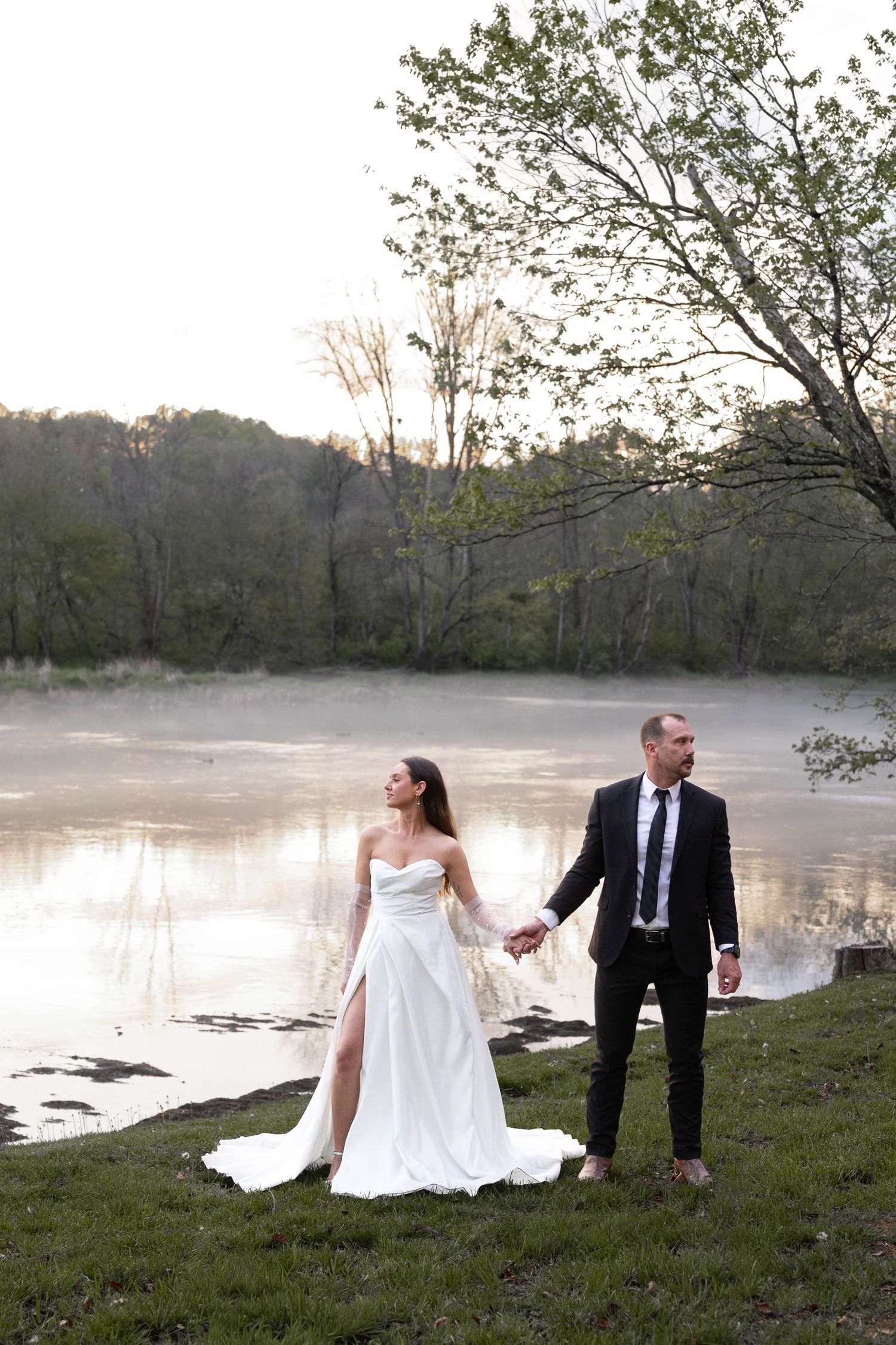 Bride in white gown and groom in black suit hold hands by a misty lake at sunset.
