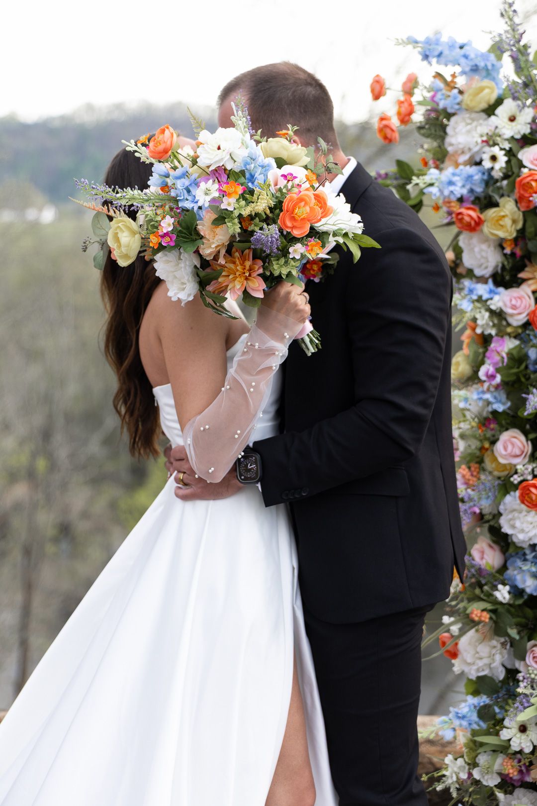 Bride and groom share a kiss behind a colorful wedding bouquet at an outdoor floral arch ceremony.