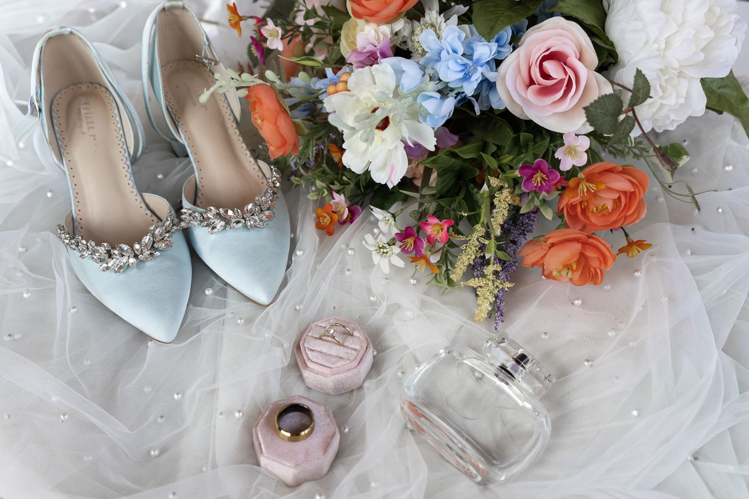 Bridal flat lay featuring silver jeweled heels, colorful bouquet, pink ring box, and perfume bottle on white tulle.