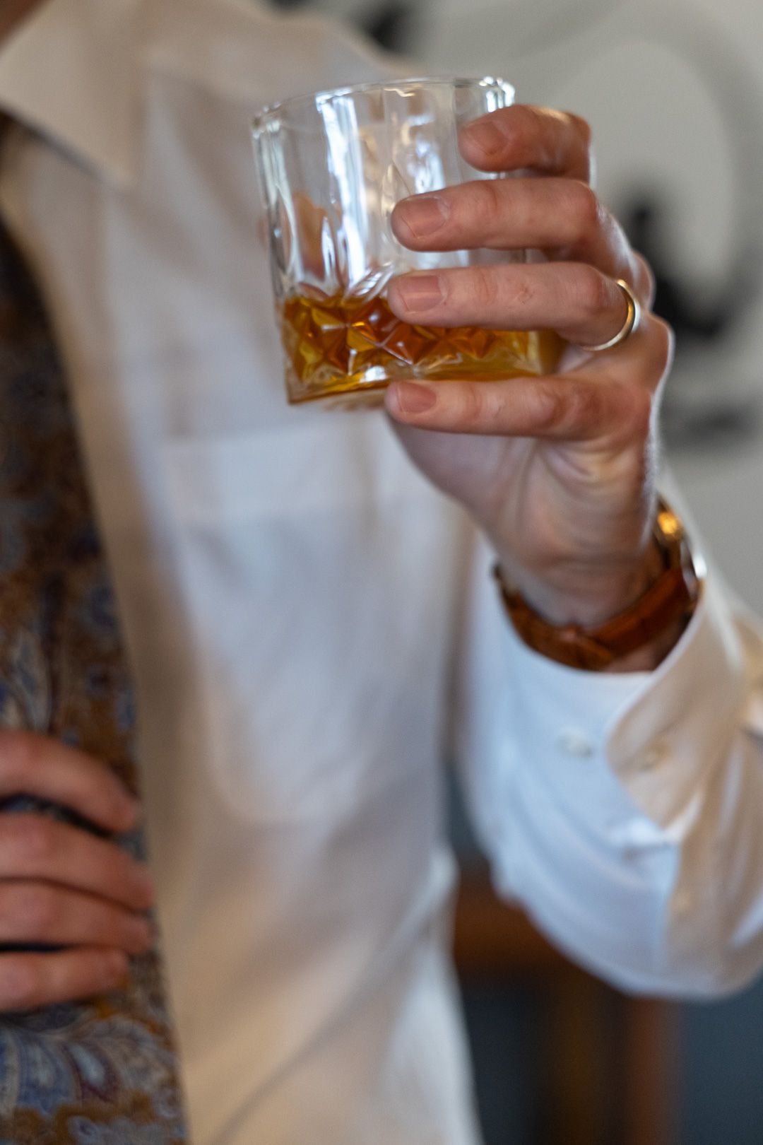 Man in white dress shirt holding a glass of whiskey on the rocks, wearing a gold ring.