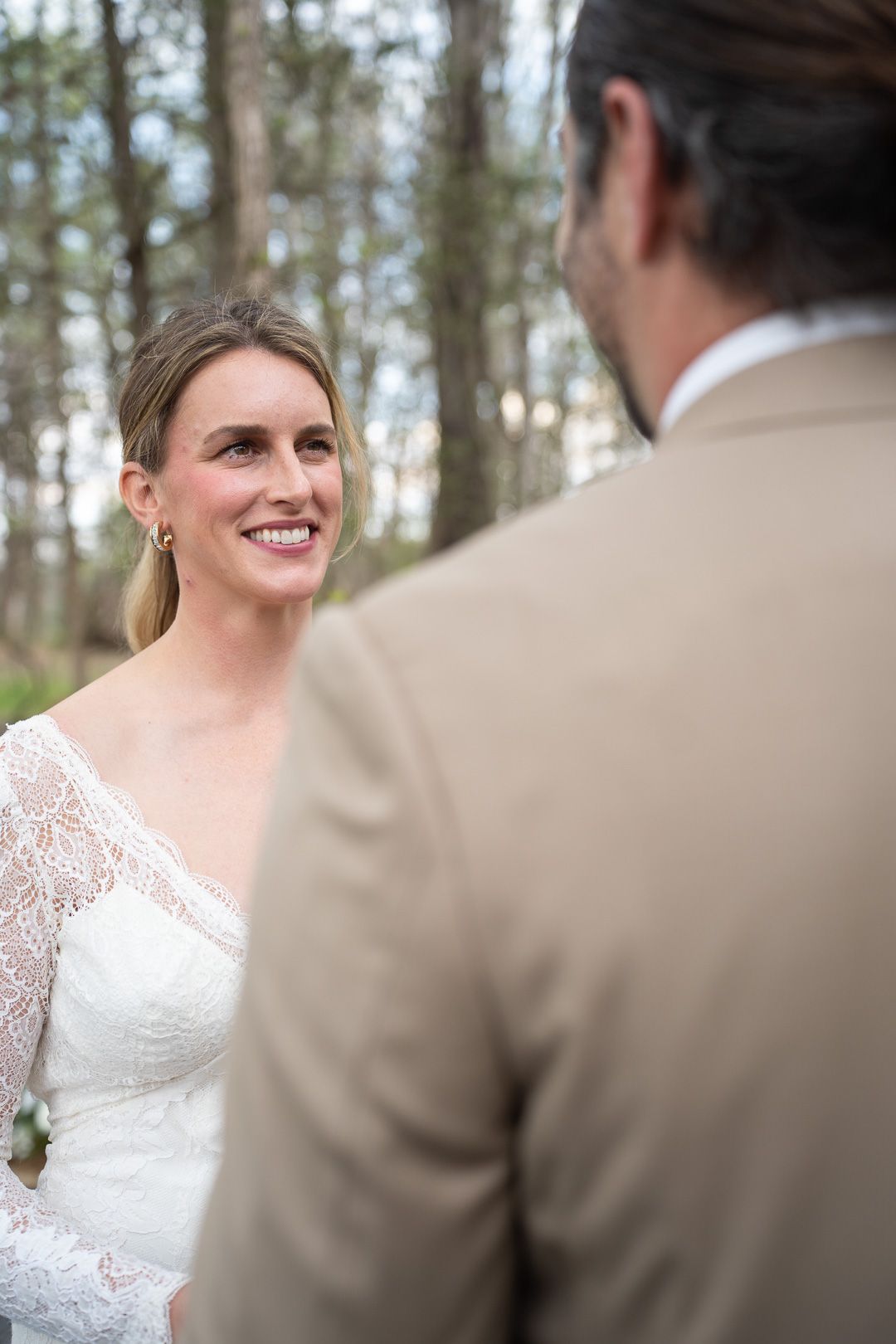 Smiling bride in white lace long-sleeve wedding dress exchanges vows with groom in tan suit during outdoor forest wedding ceremony.