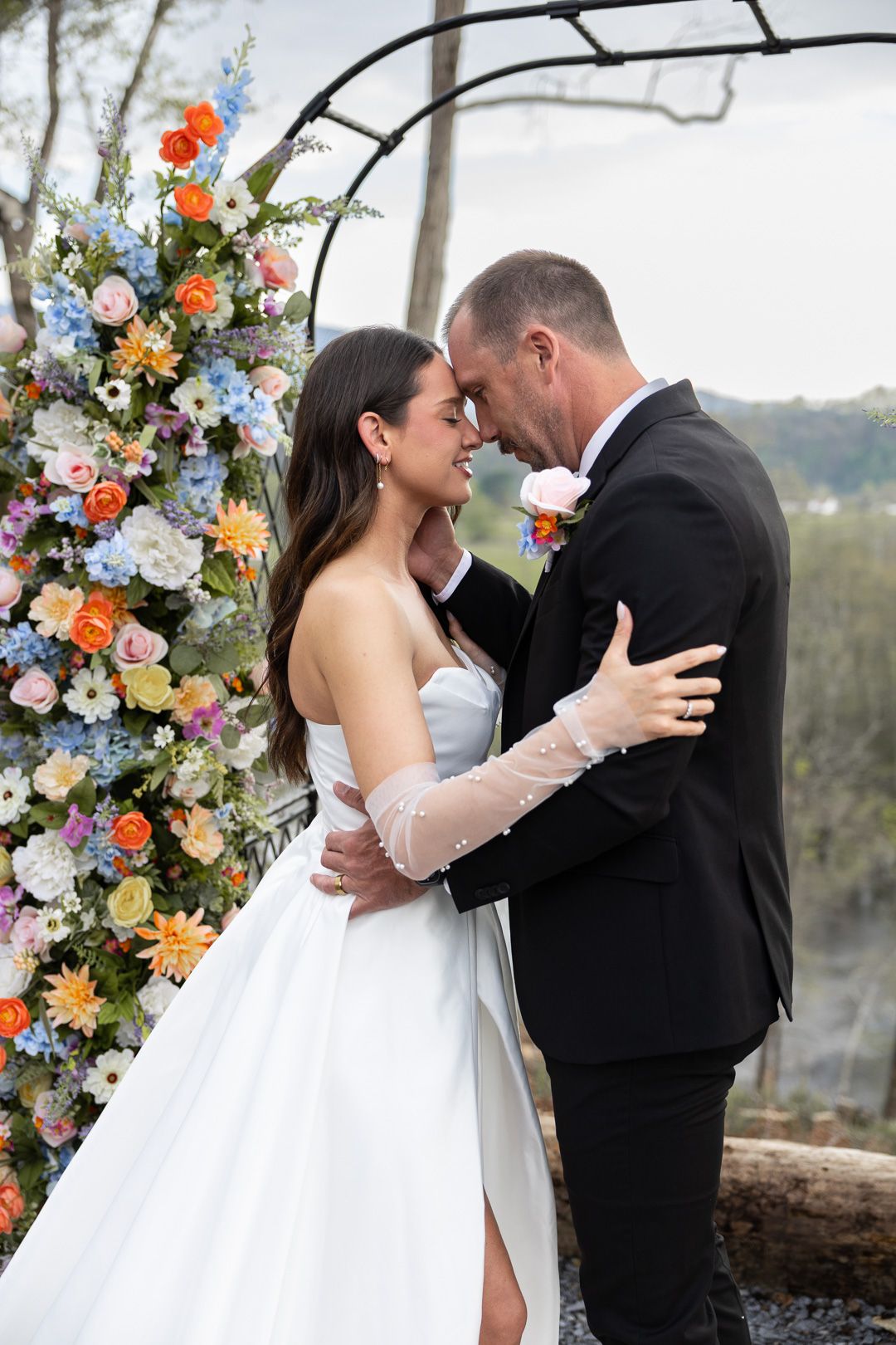 Bride and groom sharing an intimate moment under a colorful floral arch with orange, blue, and pink blooms.