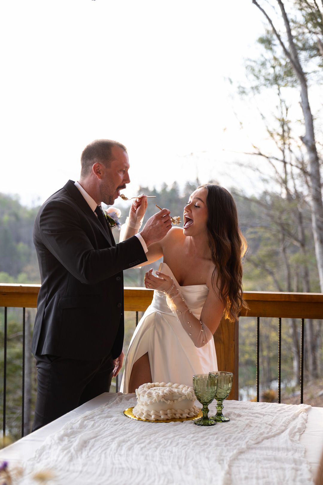 Bride and groom laugh while feeding each other wedding cake on a sunlit outdoor deck.