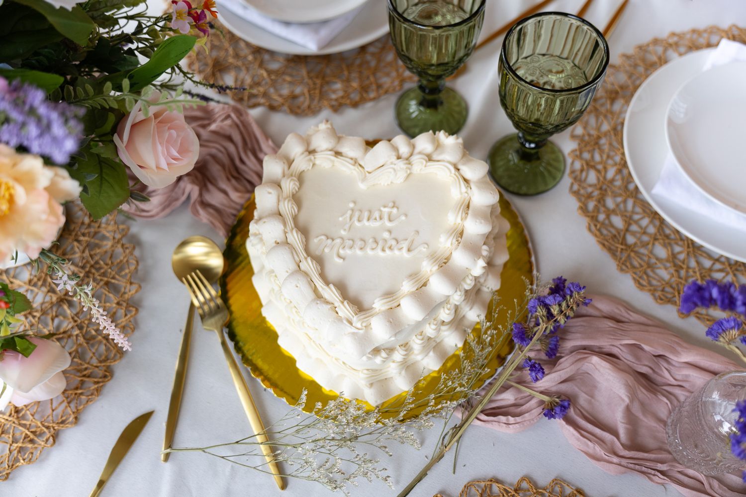 Heart-shaped white wedding cake with piped frosting reading just married on a gold stand with green goblets nearby.