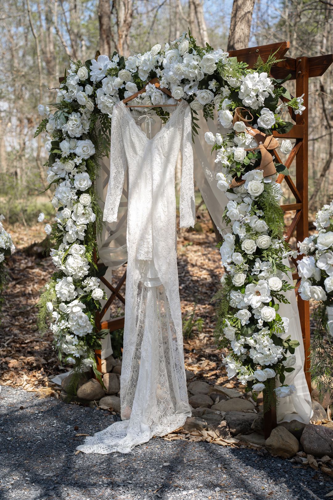 Elegant lace wedding dress hanging on a wooden arch adorned with white roses and greenery outdoors.