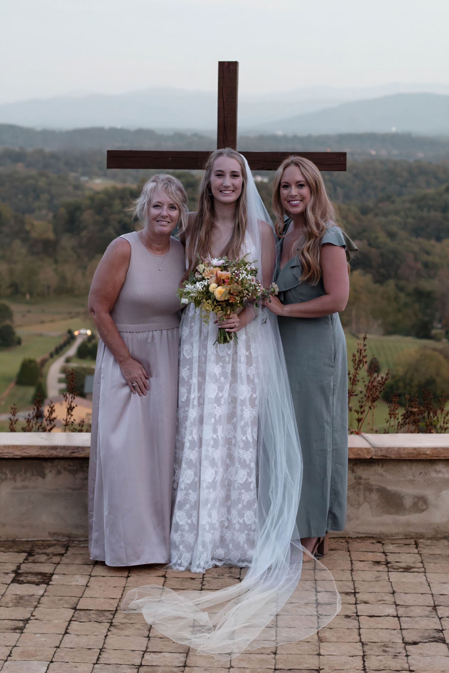 Bride in lace gown with bouquet poses with two women in maxi dresses before a cross and mountain backdrop.