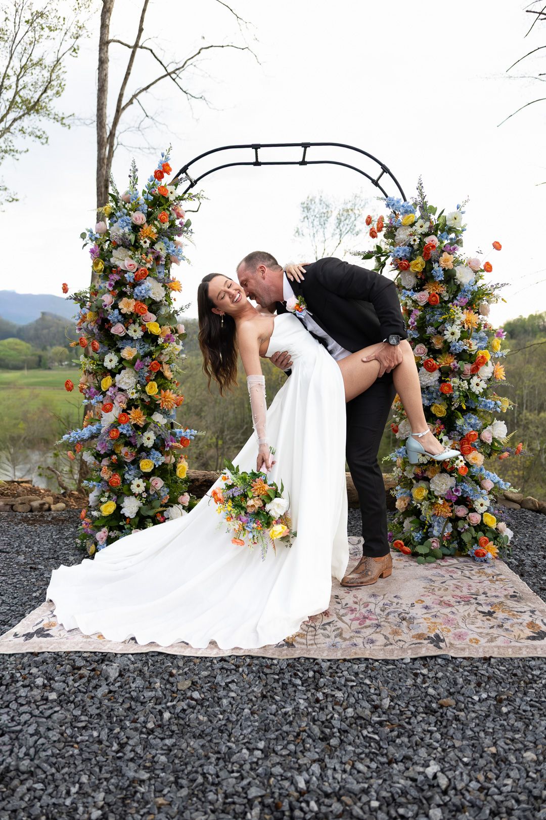 Groom dipping bride in front of a colorful floral arch outdoors with mountain views at their wedding ceremony.