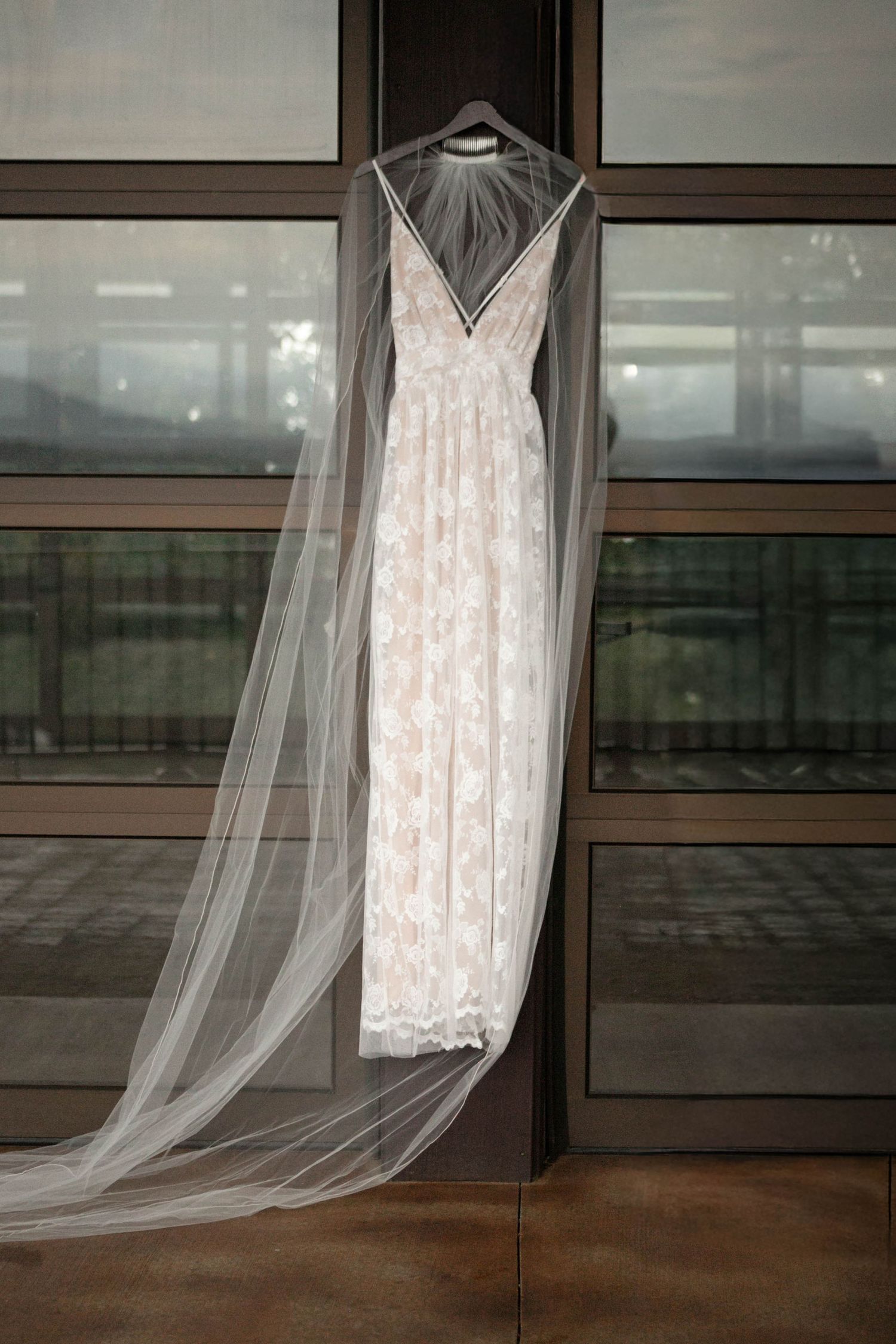 Elegant lace wedding dress with cathedral veil hanging on a door frame against large windows.