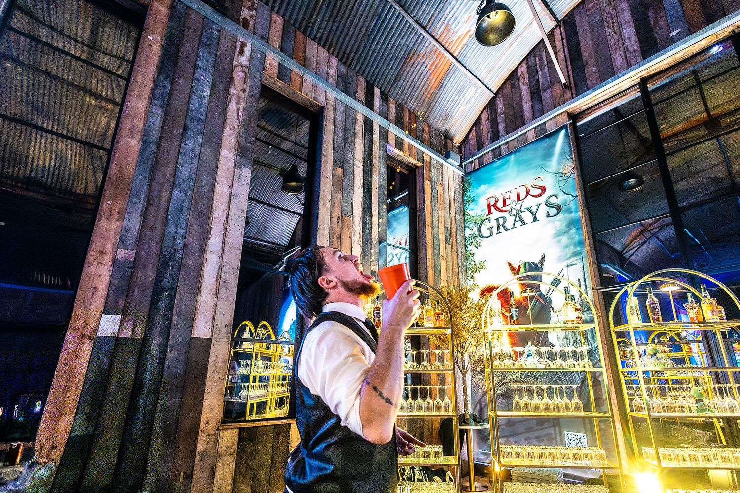 A bartender drinks from a glass at a rustic bar with a Reds vs Grays banner in an industrial venue.