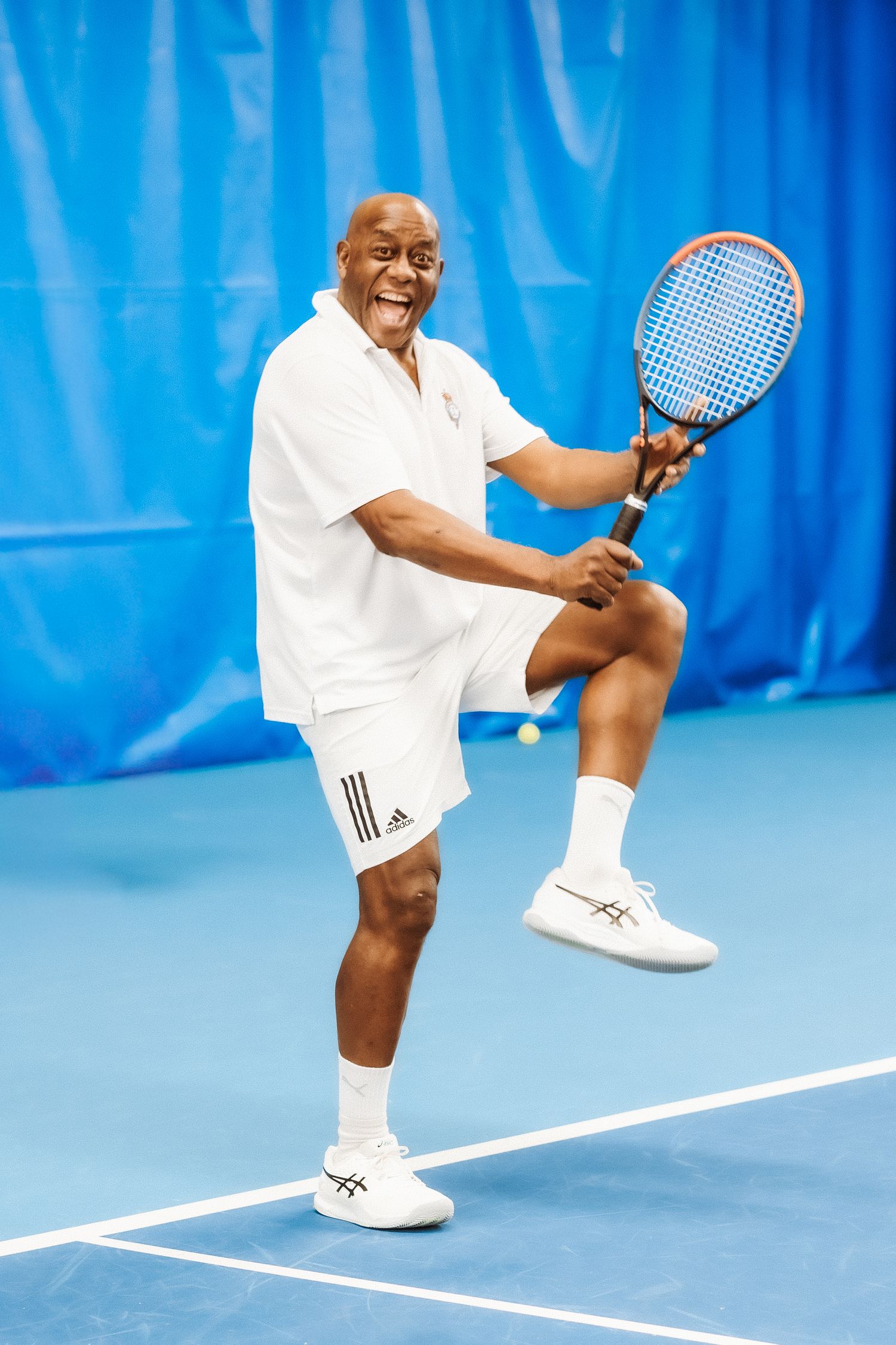 Smiling man in white Adidas tennis outfit playing tennis on an indoor blue court, mid-swing with racket.