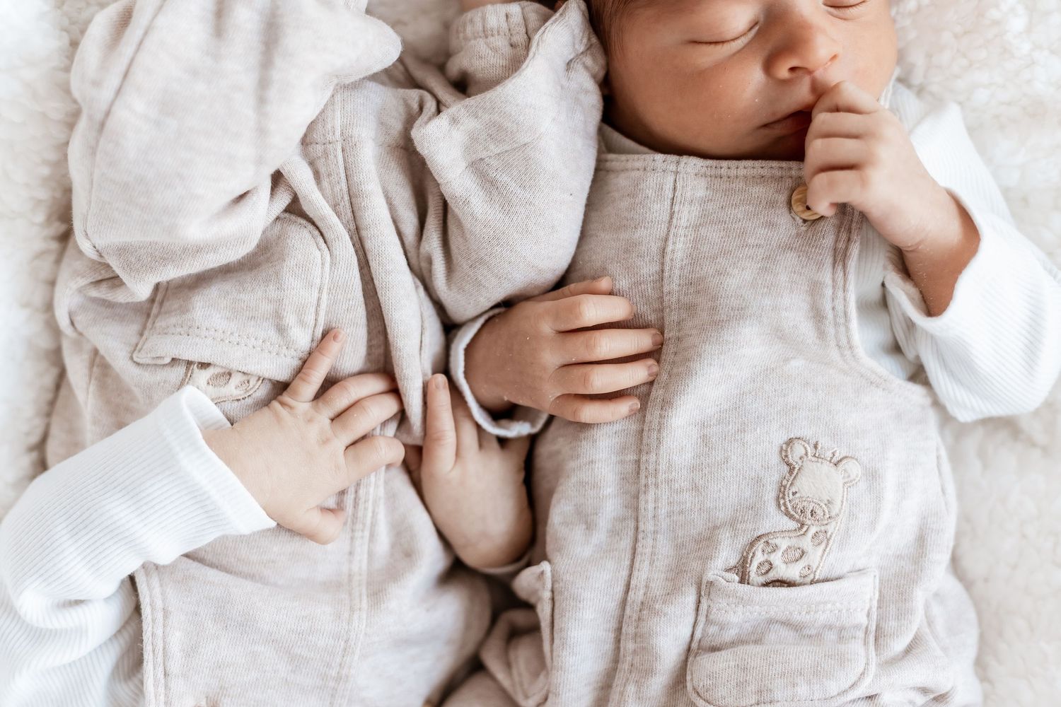 Newborn twins resting side by side on soft white bedding wearing matching gray outfits.