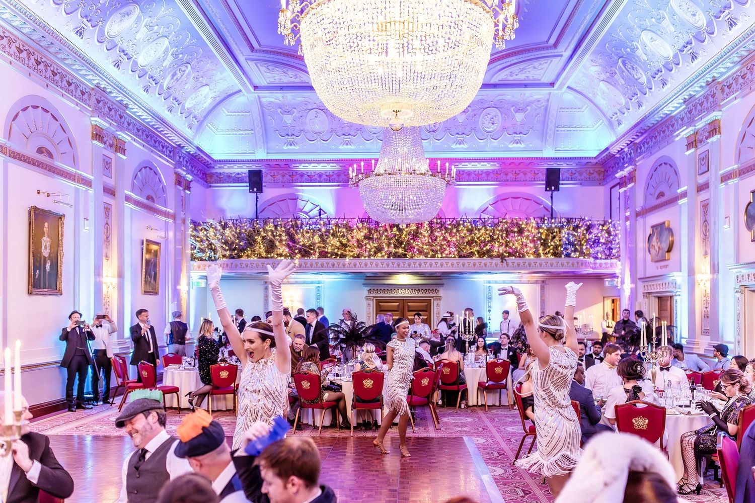 the plaisterers hall London Elegant ballroom gala with guests dancing under a grand chandelier, floral balcony, and purple ambient lighting.