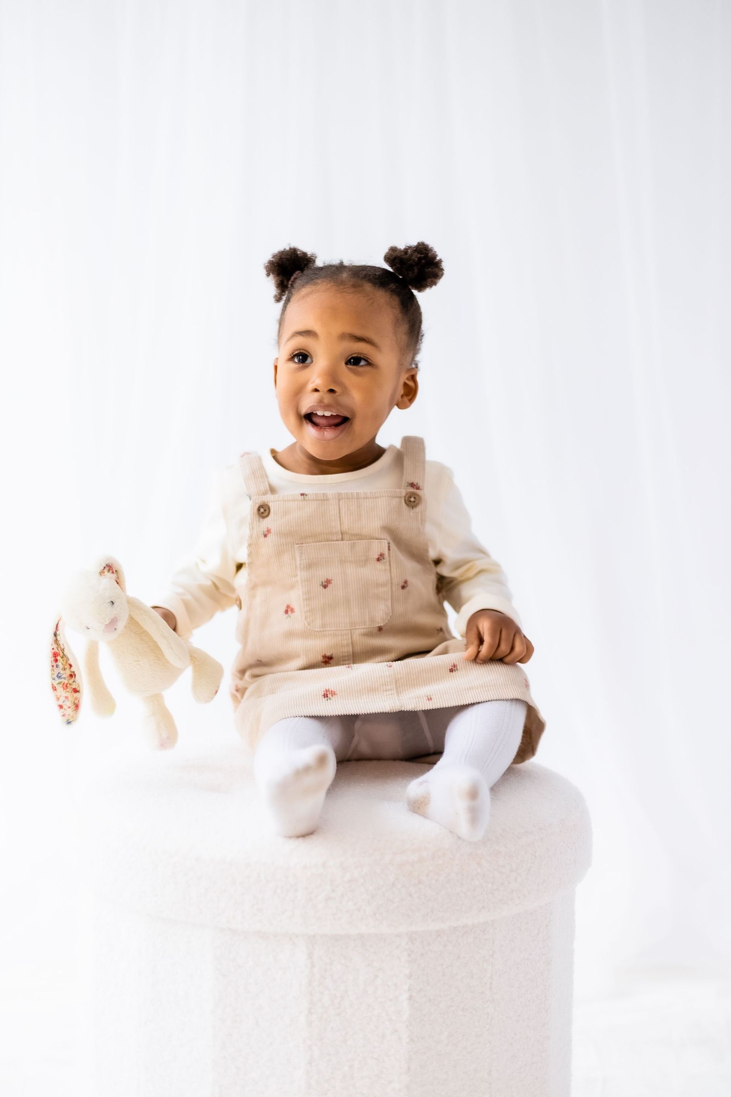 Smiling baby wearing cream-colored overalls sits on round white platform in three candid poses. Shepperton Surrey photography studio