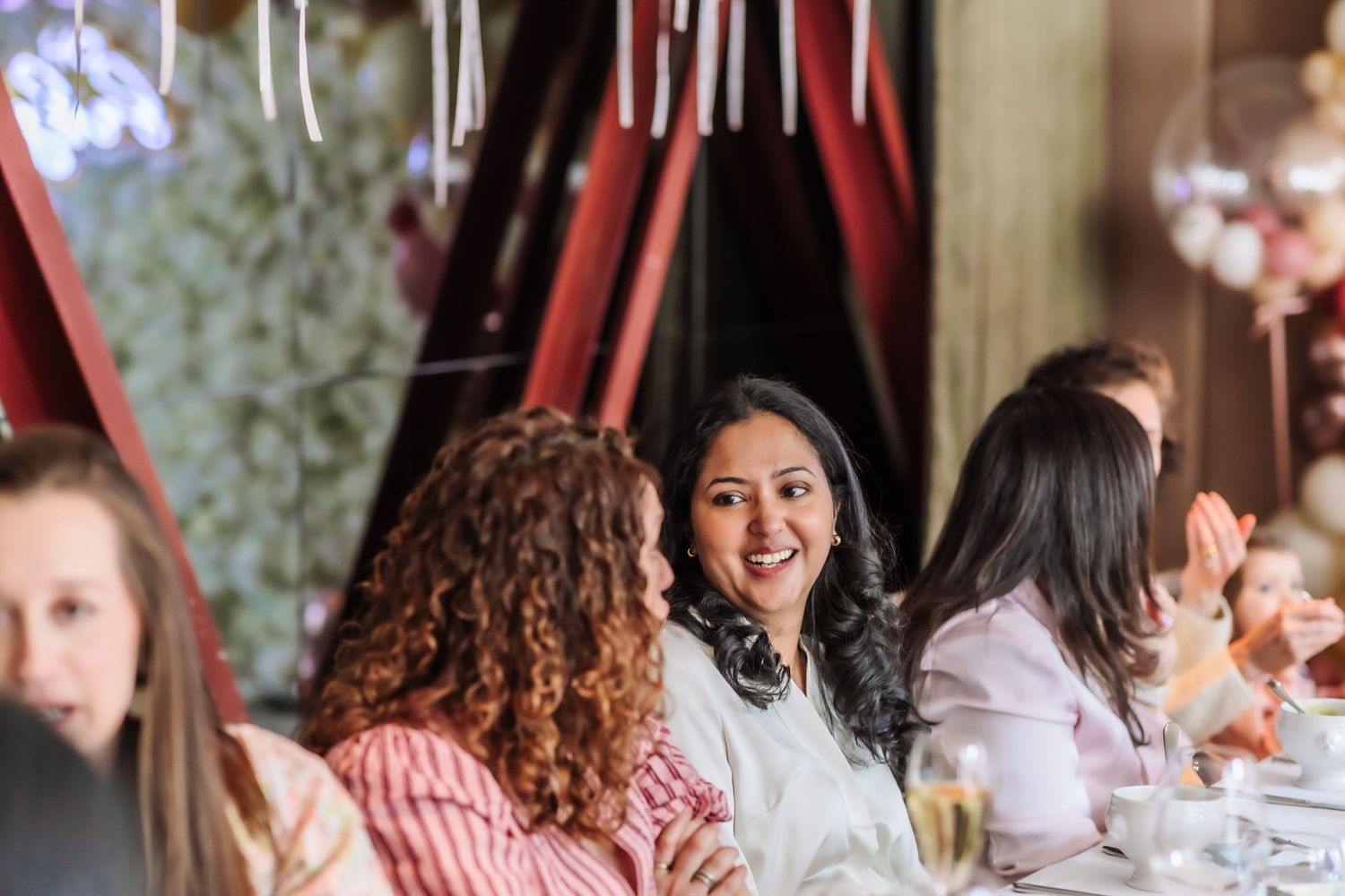 Women smiling and chatting at an elegant dinner party event with festive decorations in the background.