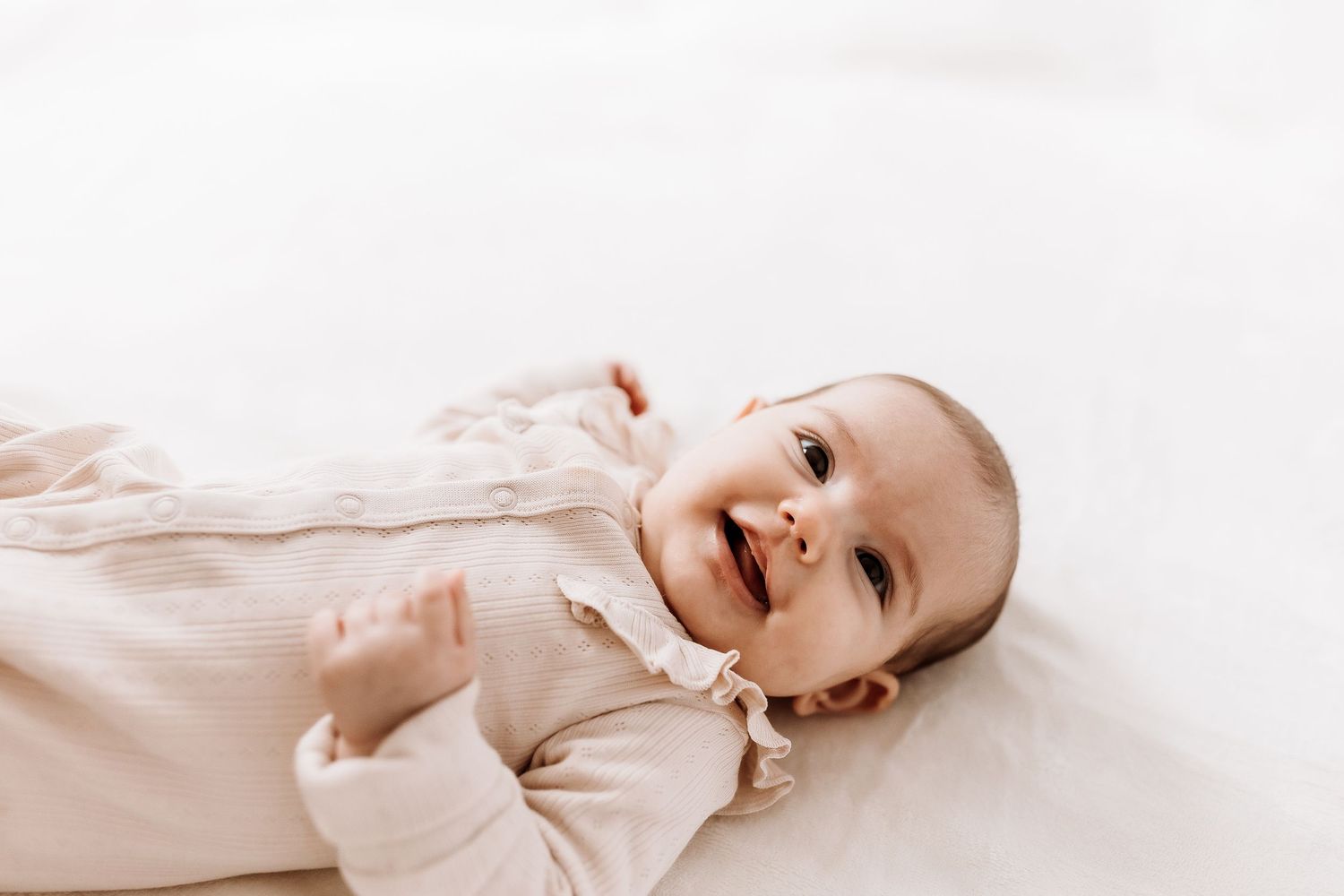 Baby wearing light clothing lies on white bedding while smiling and looking upward. candid photography baby, family Shepperton Surrey