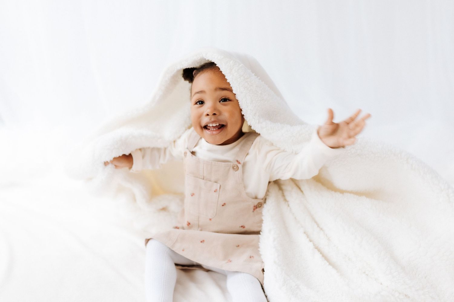A baby wrapped in white linen blankets plays and smiles while lying on a soft white surface.