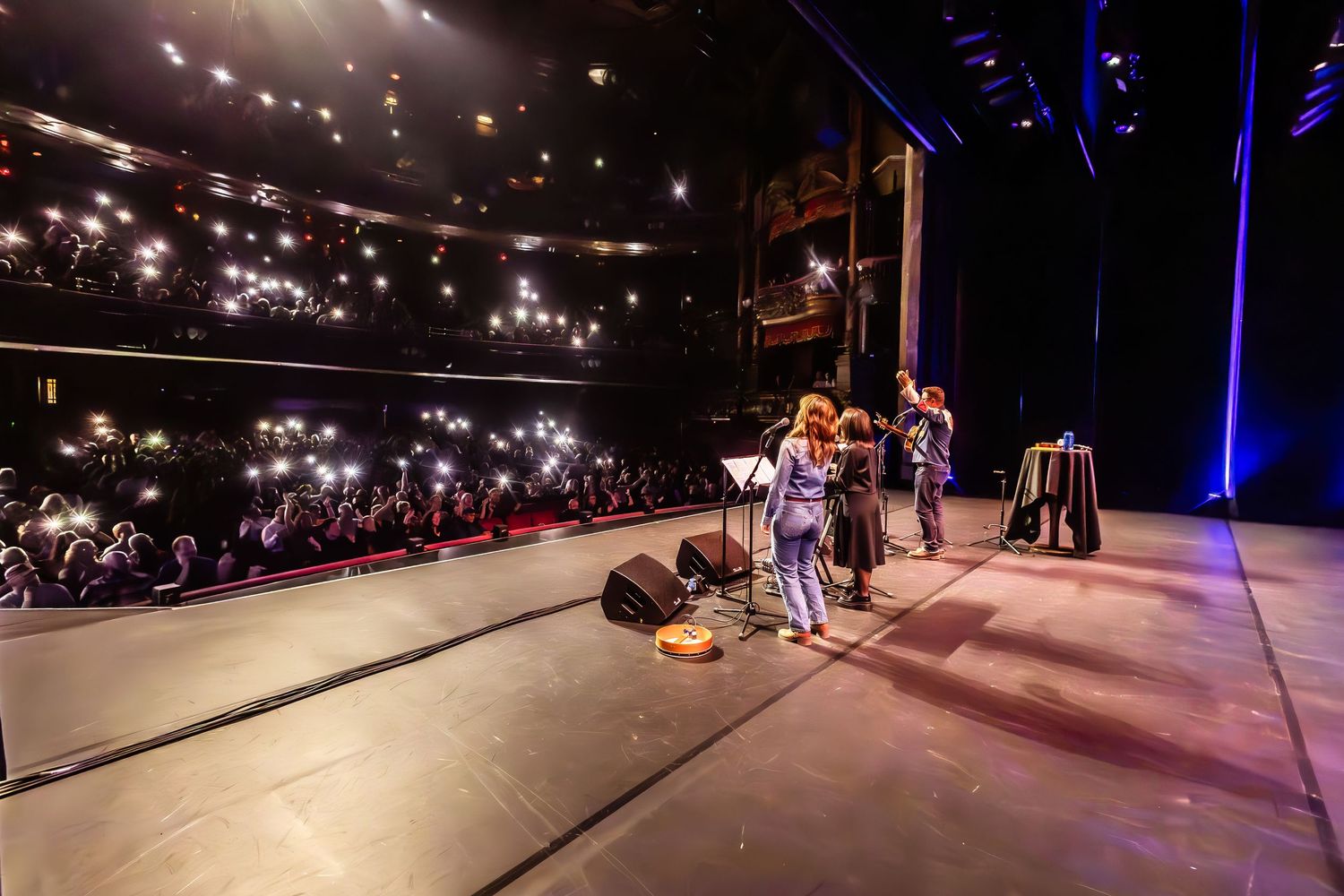 Live concert performance on stage in a grand theater with audience holding up phone lights in the dark.