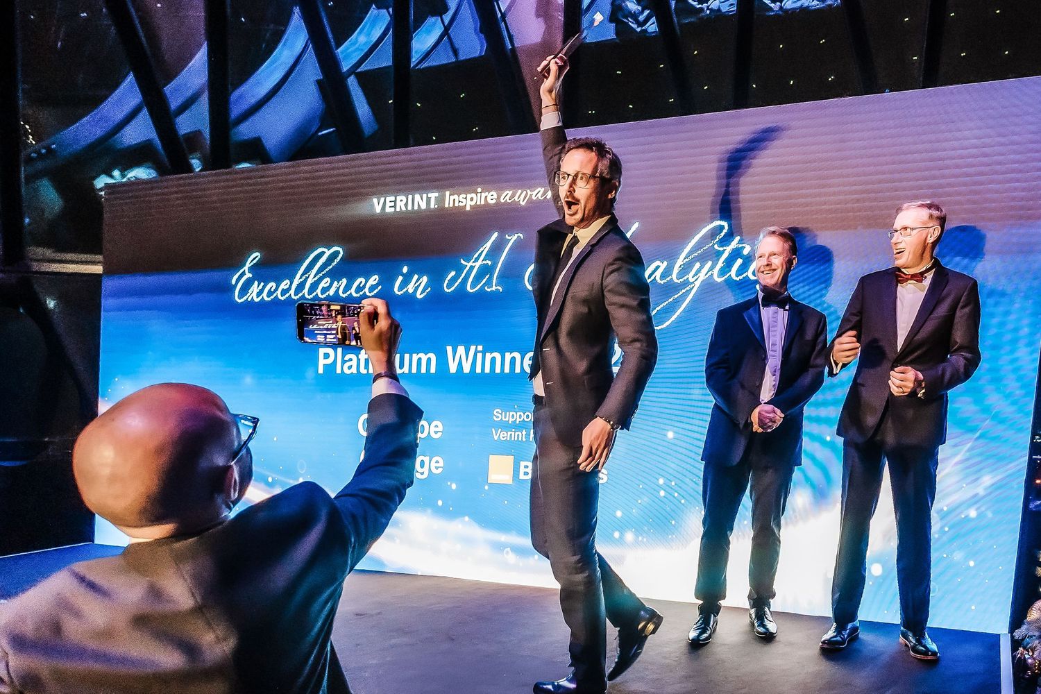 Award ceremony on stage with Verint Excellence in Analytics Platinum Winner banner, men celebrating under dramatic lighting.