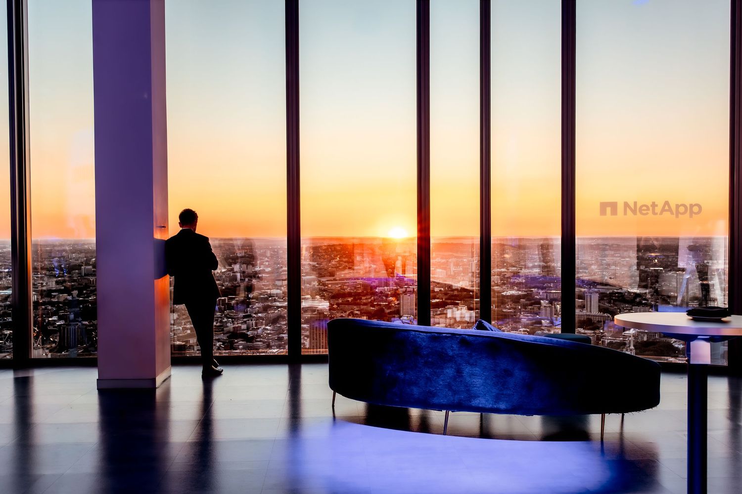 Person gazing at stunning city sunset from high-rise office with NetApp branding and modern blue sofa.