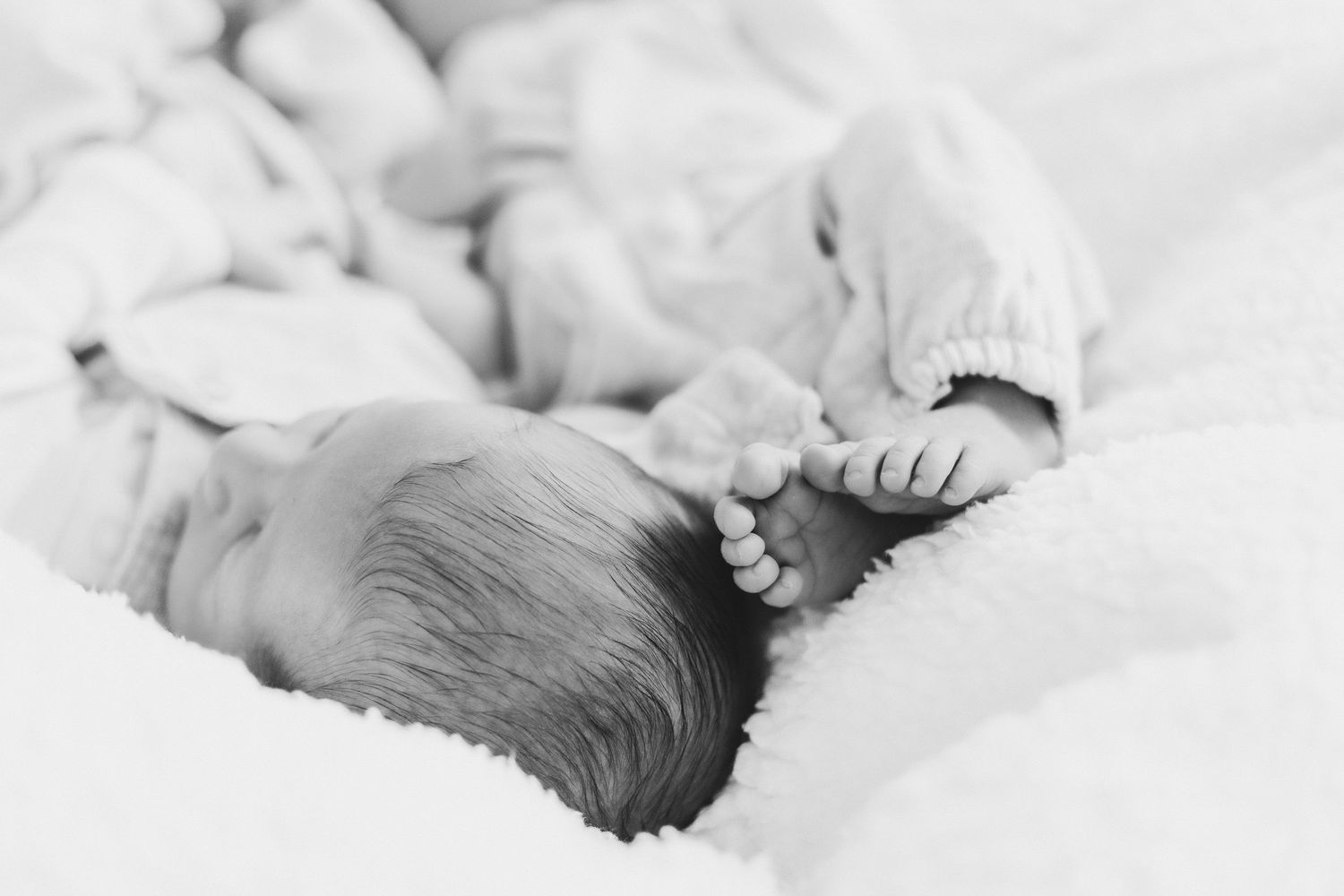 Black and white close-up of a sleeping newborn baby's head and tiny feet nestled in soft white bedding.