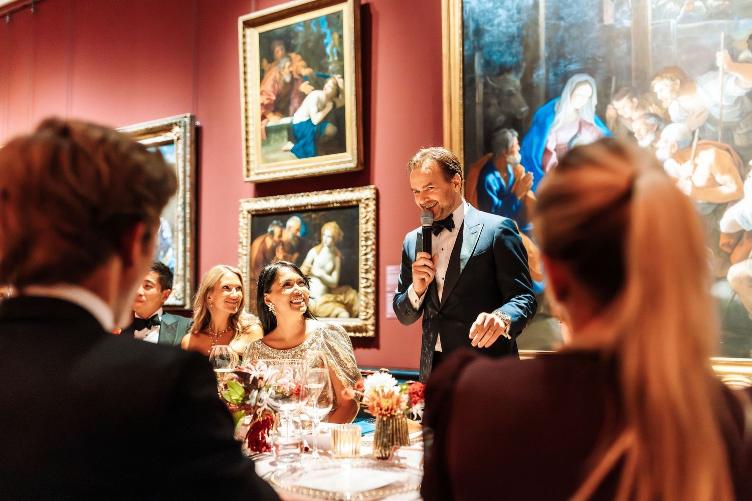 Elegant formal dinner event in a museum gallery with guests laughing as a man in a tuxedo gives a toast.