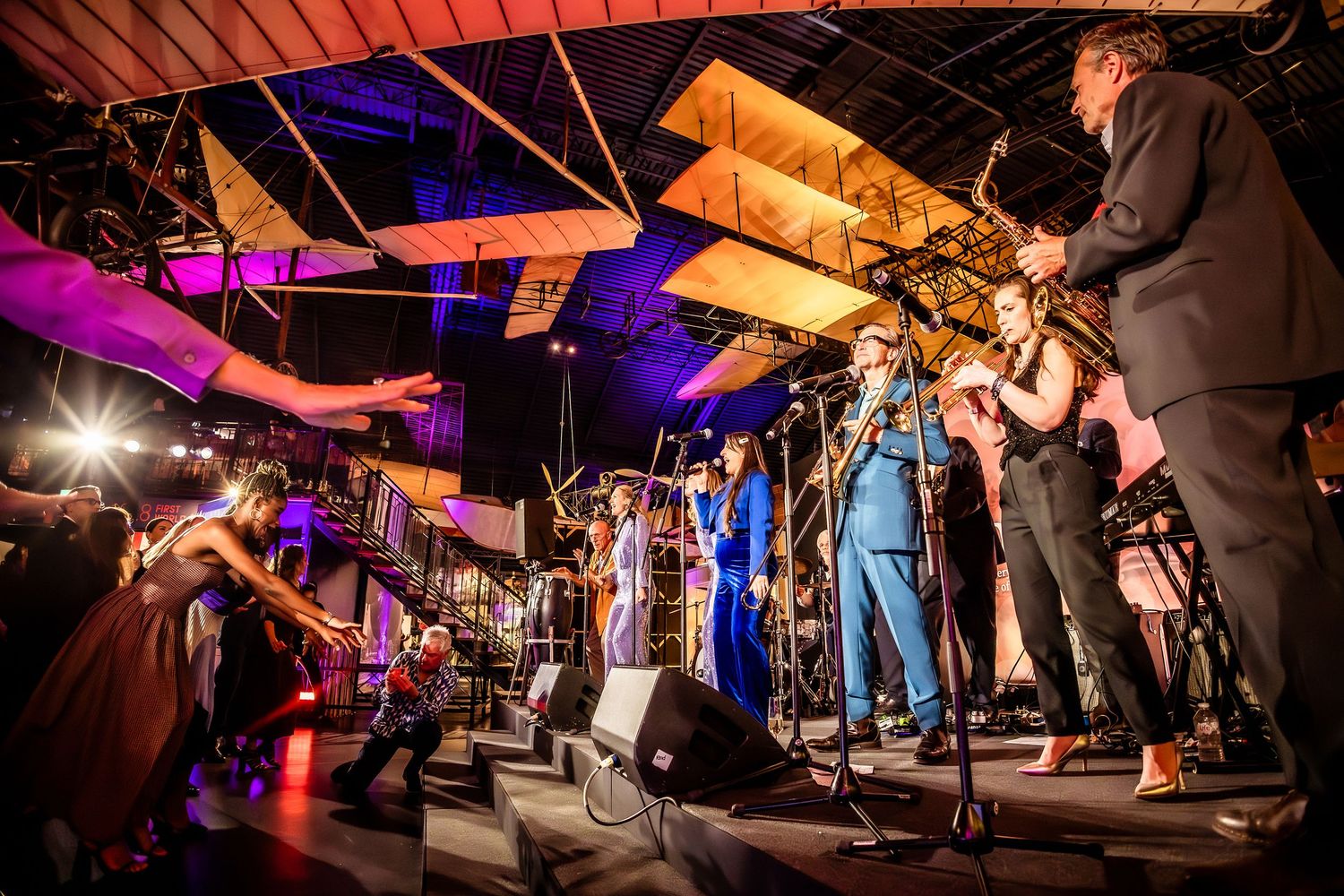 Live jazz band performing on stage with saxophones and brass instruments under vintage aircraft in a museum venue.