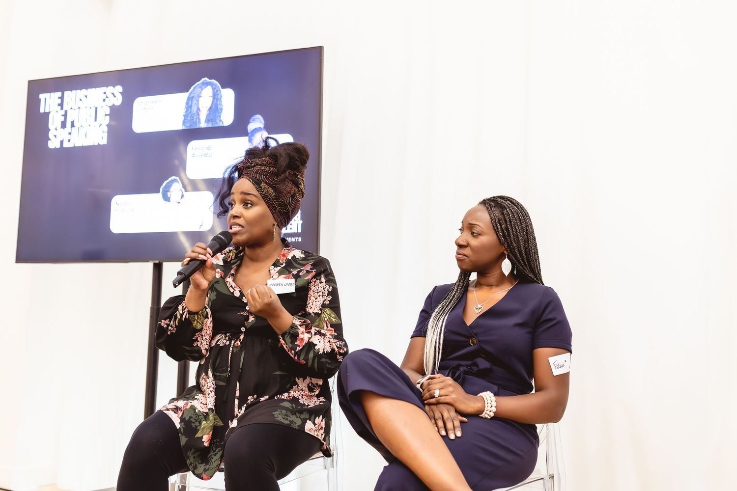 Two women on stage at a business panel event, one speaking into a microphone, seated in front of a presentation screen.