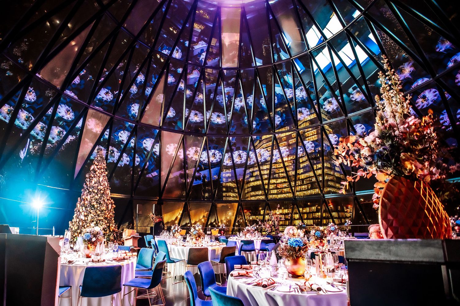 Elegant Christmas gala dinner in a stunning domed venue with festive trees, blue chairs, and colorful stained glass windows.