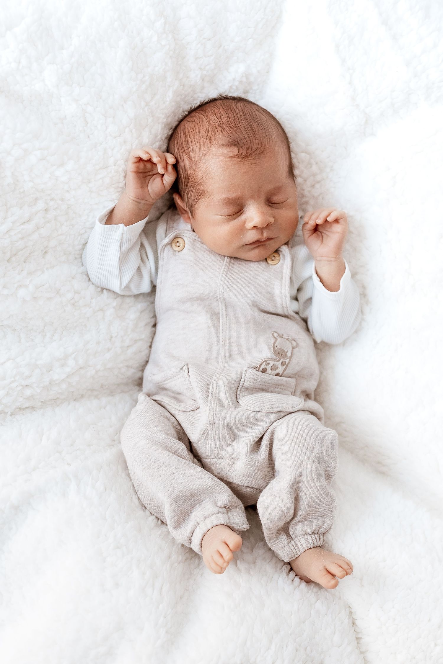 Peaceful newborn wearing light beige outfit sleeps on white textured blanket.