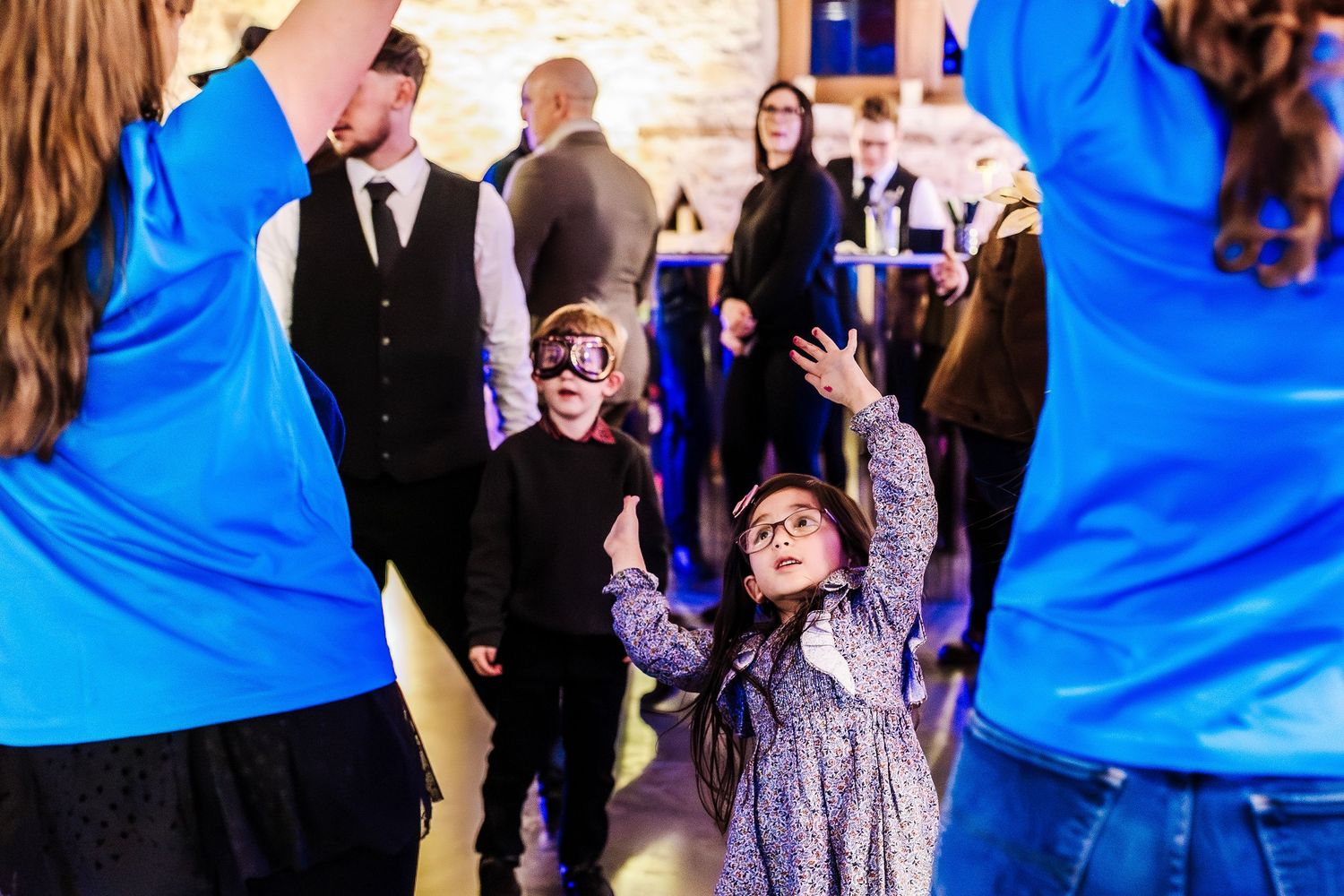 Children in costumes dance joyfully at a festive indoor party event with adults in formal attire in the background.