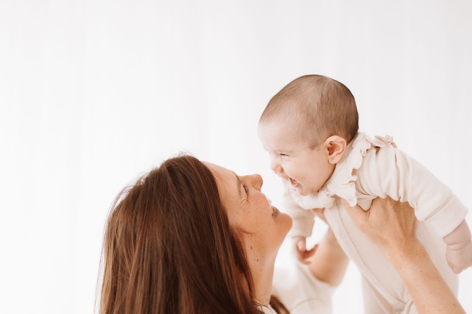 A mother holds up a baby against a white background in a tender moment of connection and play.
