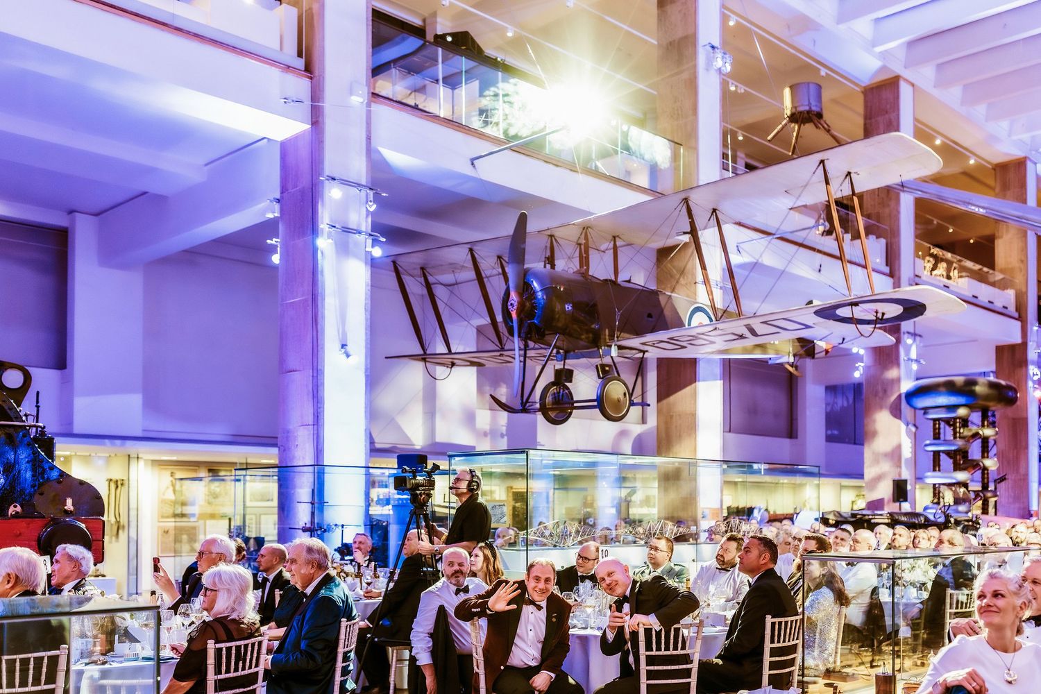 Elegant gala dinner event at the science museum with vintage aircraft suspended from ceiling and guests seated at white-draped tables.