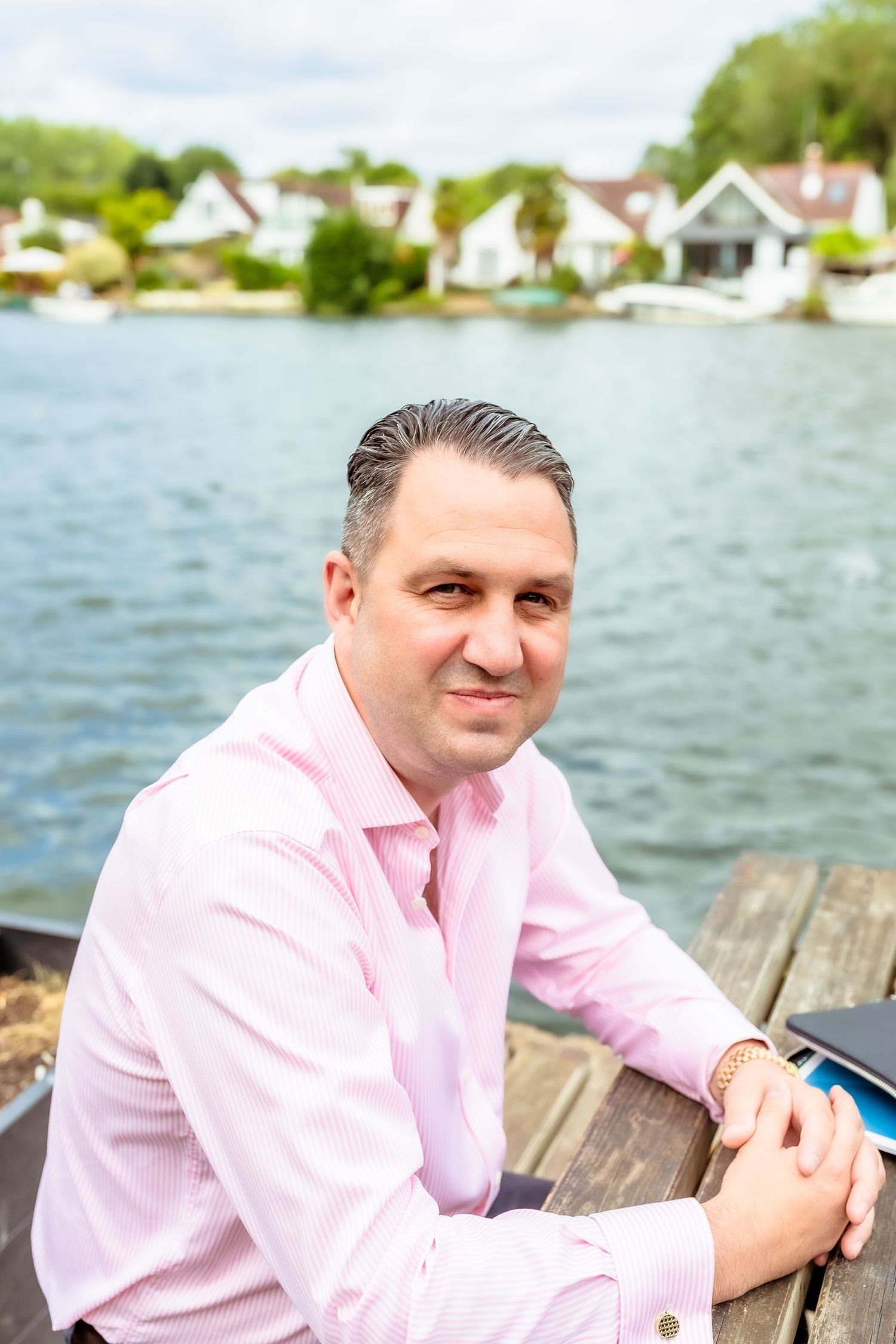 Person in pink button-down shirt sitting on wooden dock by lake with waterfront homes in background.