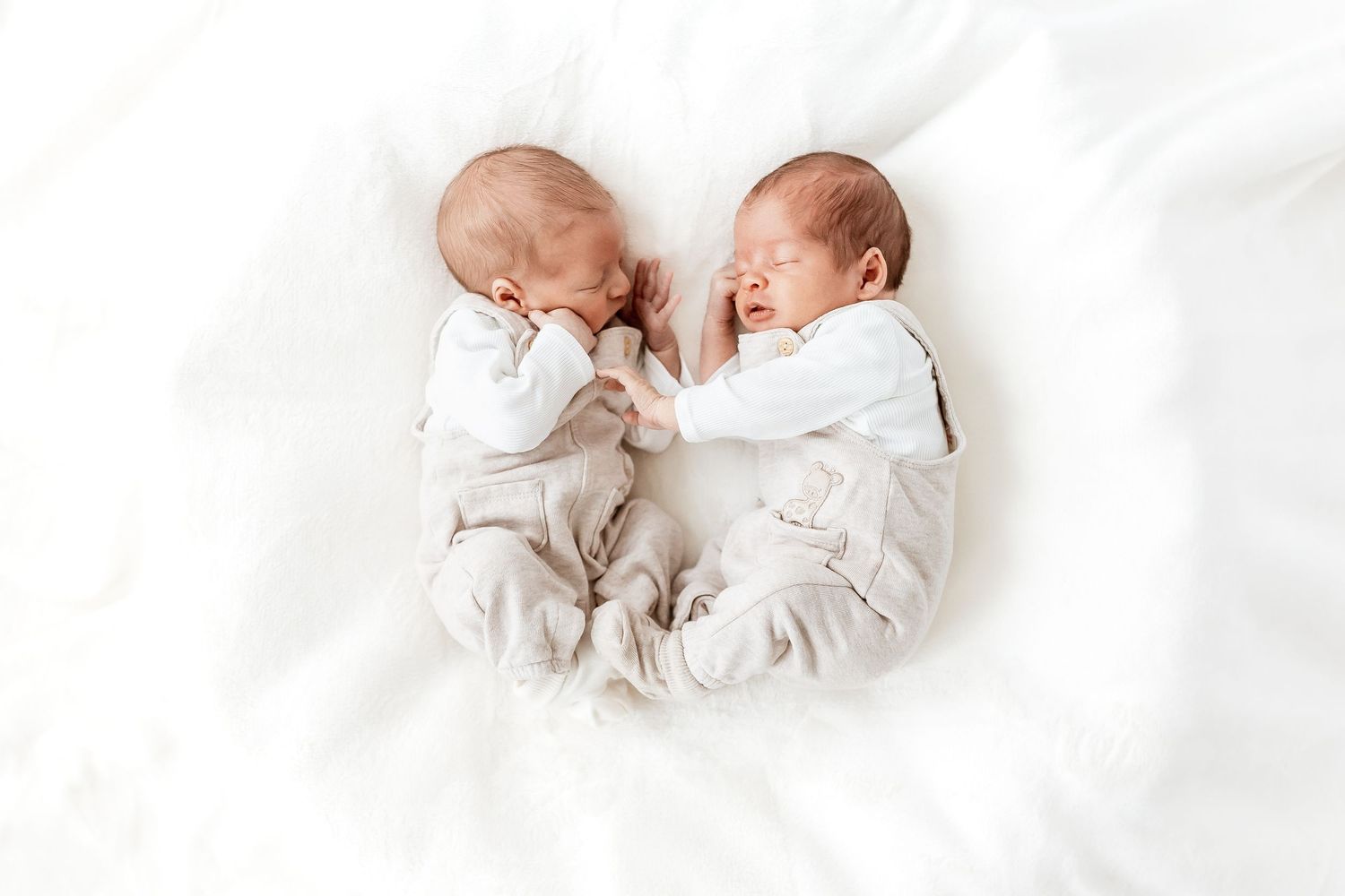 Twin newborns sleeping peacefully together on white bedding in matching outfits.