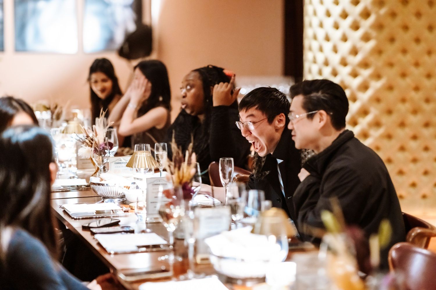 Diverse group of guests dining at a long restaurant table with elegant decor and glassware.