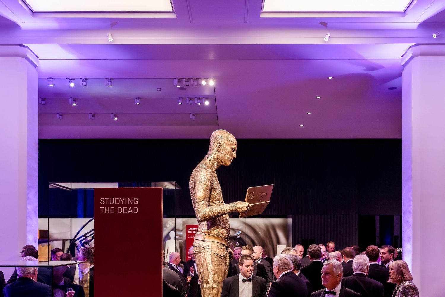 A tattooed presenter speaks at a podium labeled 'Studying the Dead' during a formal gala event.