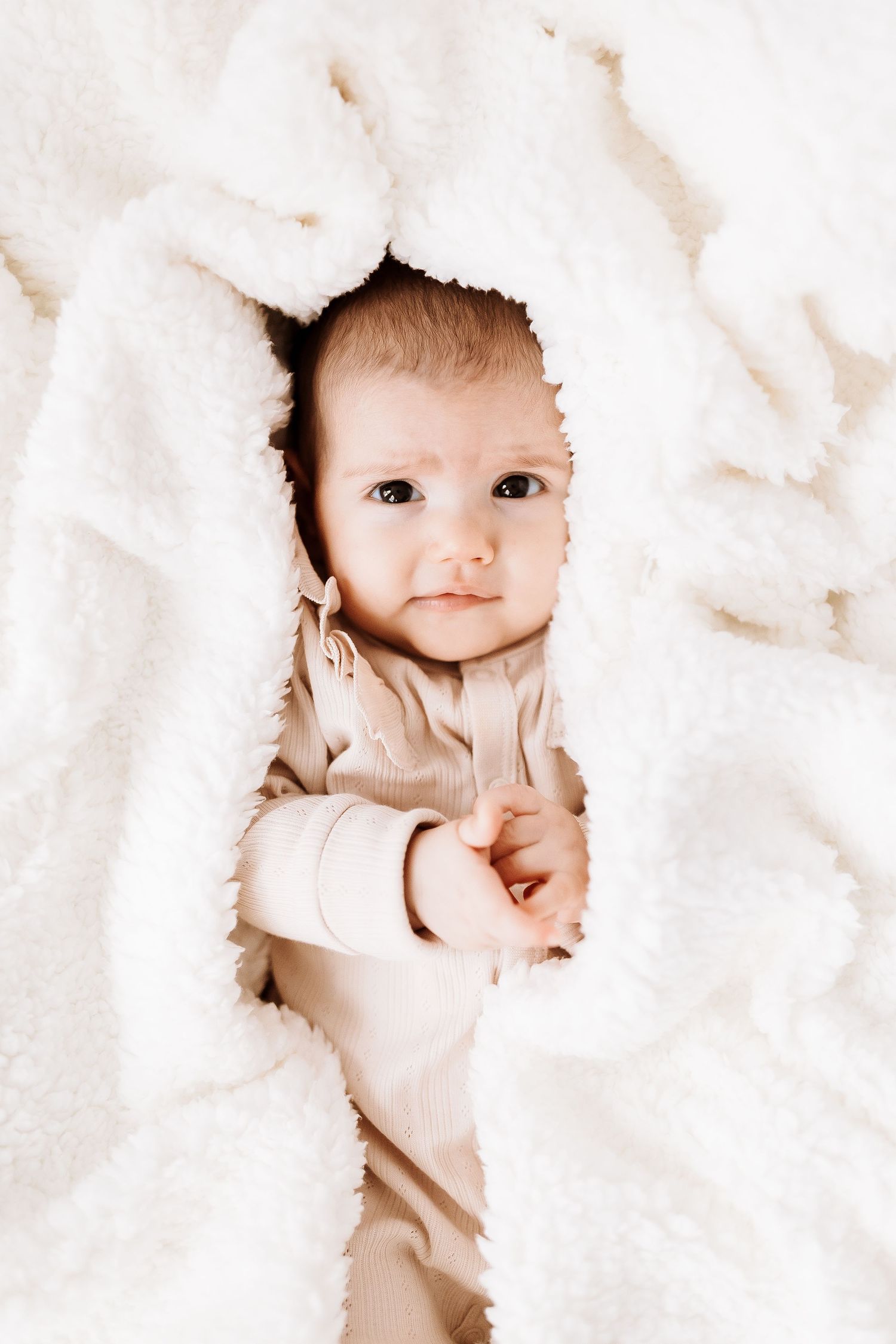 A baby peeks playfully through soft white blankets creating a cozy framed portrait effect. candid photography baby, family Shepperton Surrey