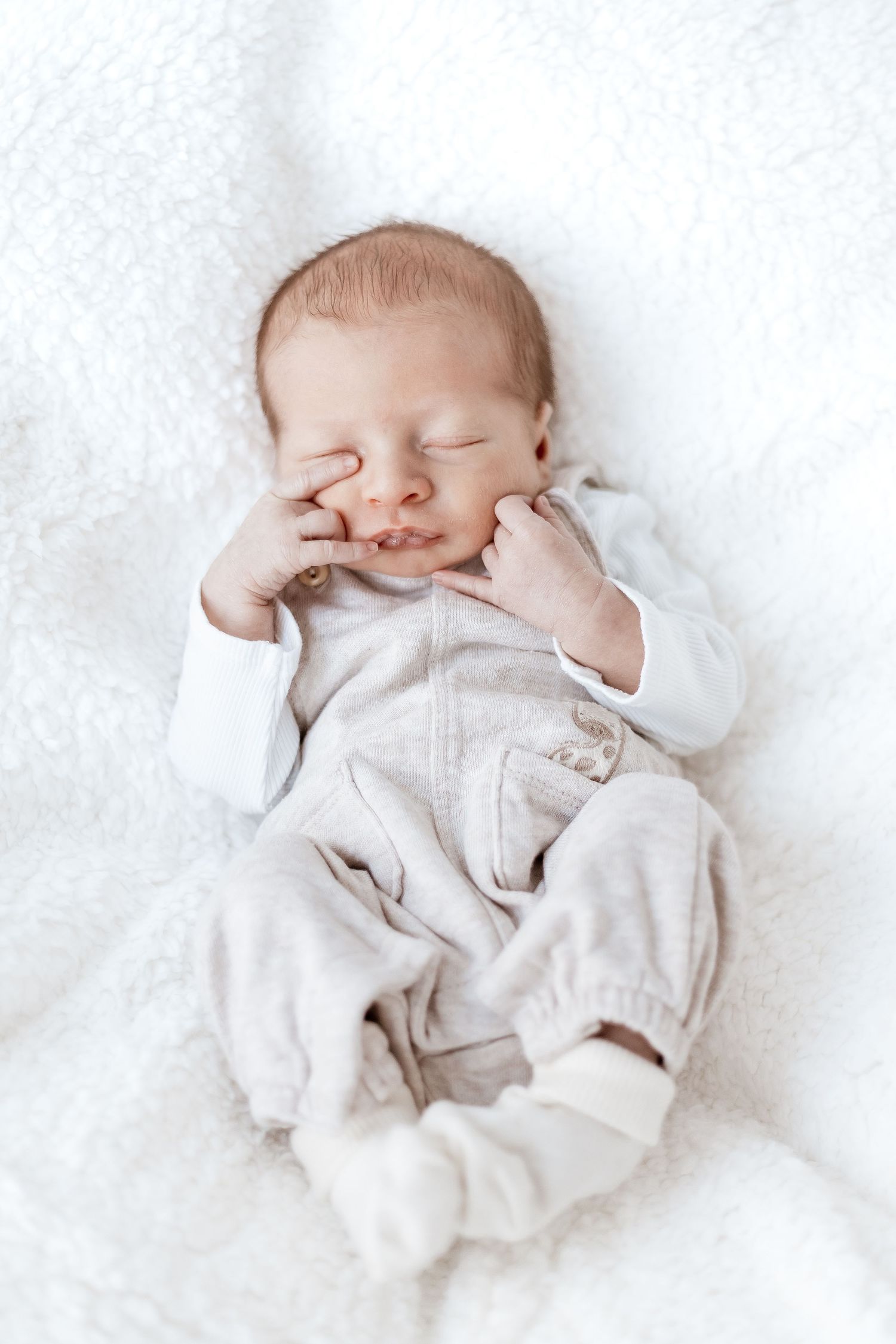 Sleeping newborn in soft white clothing rests peacefully on plush white bedding.