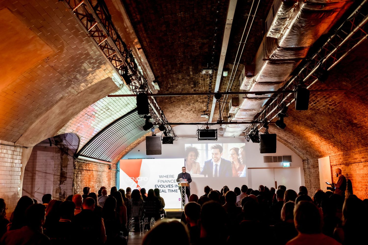 Guests gather in a brick-vaulted underground venue for a formal event with a large presentation screen.