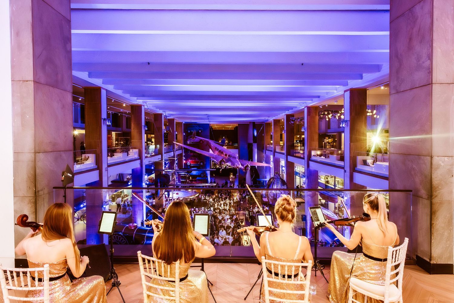 String quartet performing on a balcony overlooking an elegant event venue with purple ambient lighting.