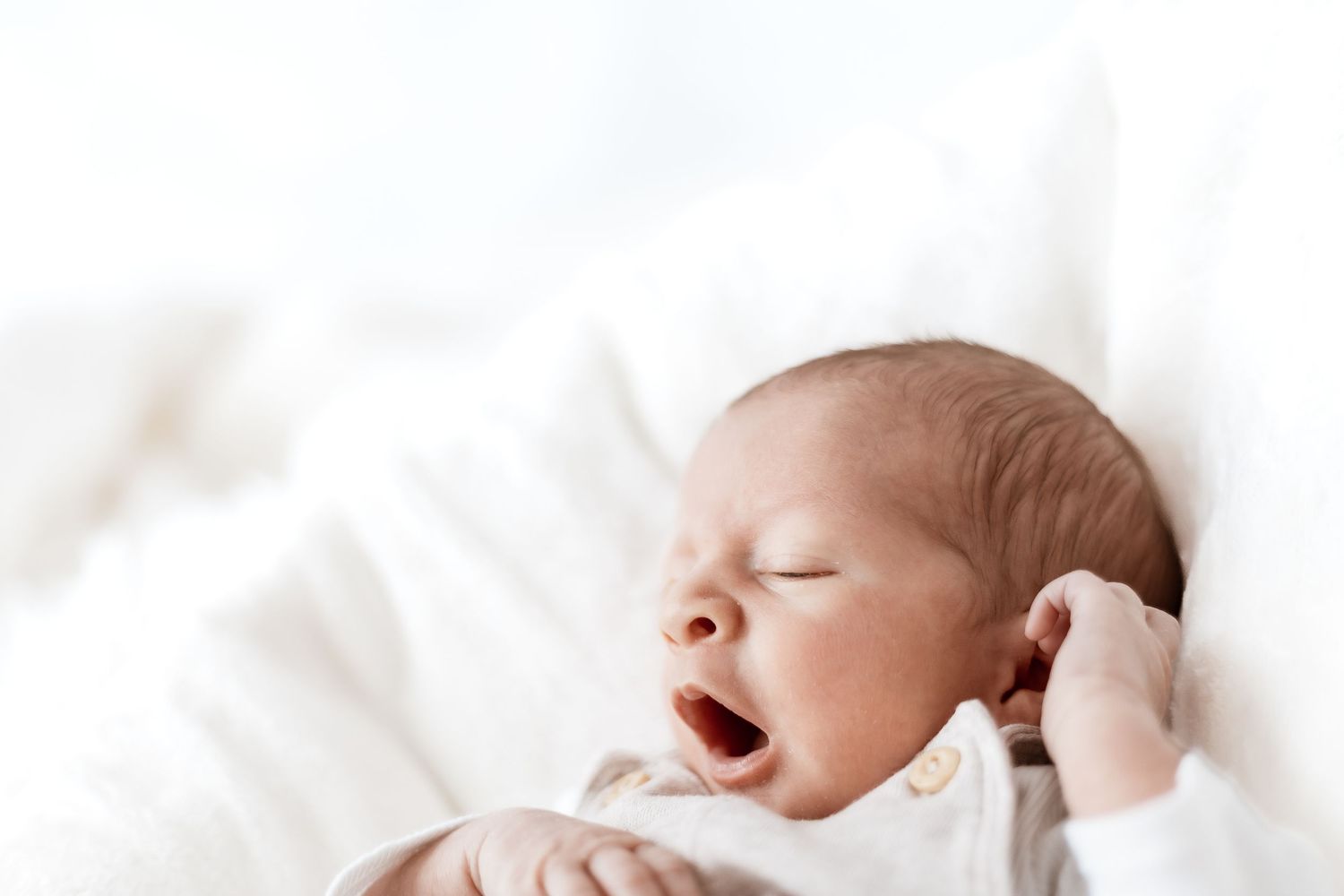 A tiny newborn baby yawns while resting on soft white bedding.