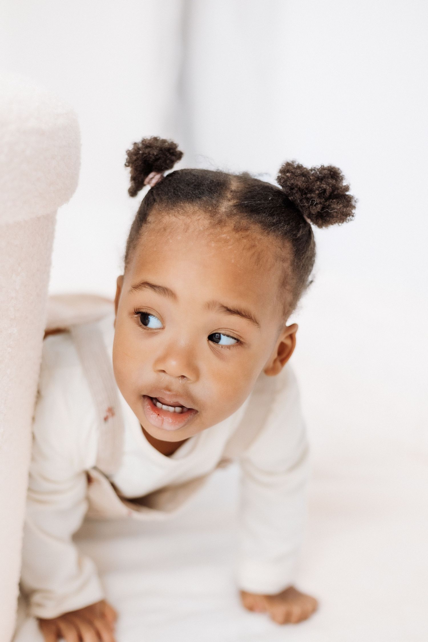 Playful child peeks through white furniture with bright smile and hair buns. Shepperton Surrey photography studio portrait baby session
