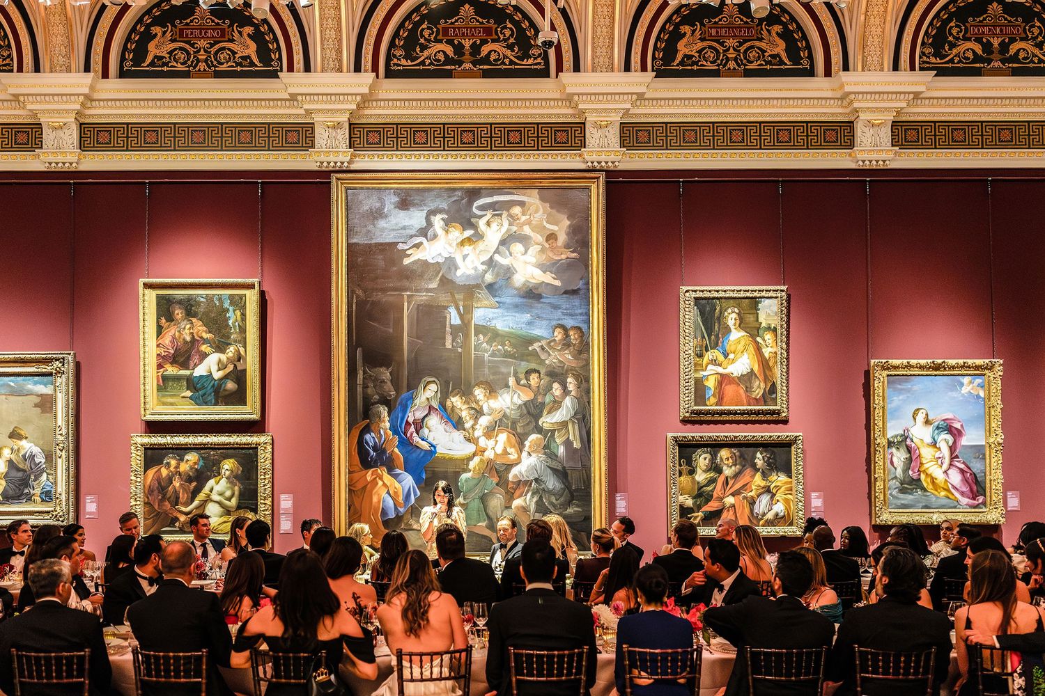 Elegant gala dinner in a grand museum gallery with classical paintings on red walls and formally dressed guests.