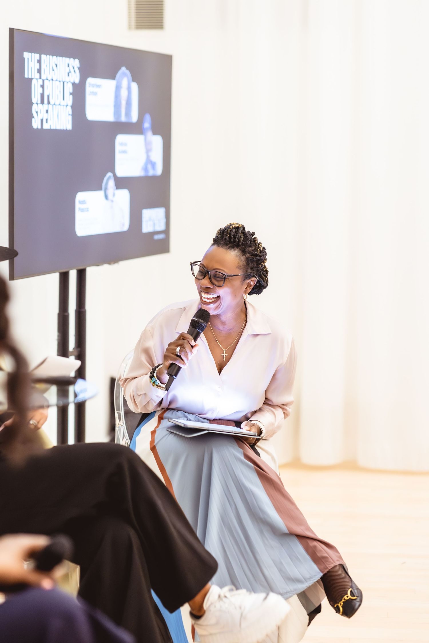 A smiling woman speaks into a microphone at a public speaking business event, holding notes while seated on stage.